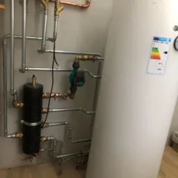 Instalacja grzewcza z białym zasobnikiem ciepłej wody, widoczne rury miedziane i zawory, zamontowana na szarej ścianie w pomieszczeniu z drewnianą podłogą.