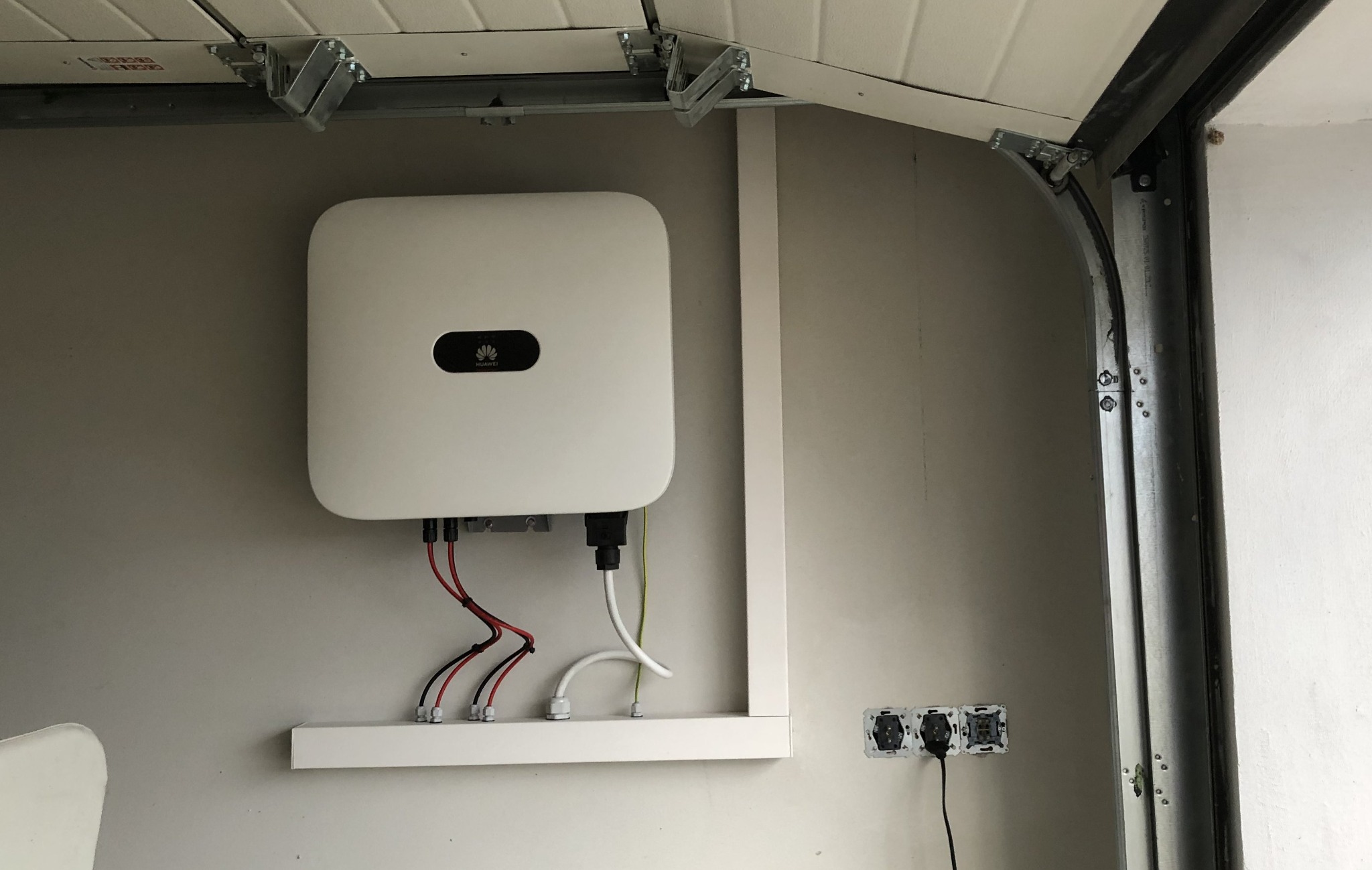 Biały falownik fotowoltaiczny Huawei zamontowany na szarej ścianie w garażu, widoczne przewody, listwa montażowa i gniazdka elektryczne.