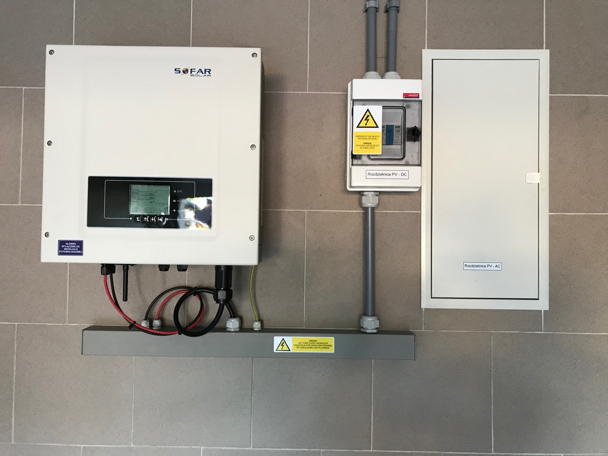 Biały falownik SOFAR Solar, skrzynka rozdzielcza PV-DC z ostrzeżeniem o wysokim napięciu, oraz skrzynka rozdzielcza PV-AC zamontowane na szarej ścianie z płytek.