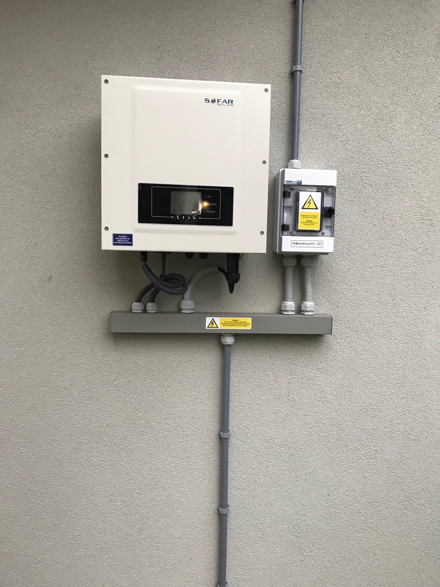 Biały falownik Sofar Solar zamontowany na szarej ścianie zewnętrznej budynku, z widoczną skrzynką przyłączeniową DC i przewodami w szarych rurkach instalacyjnych.