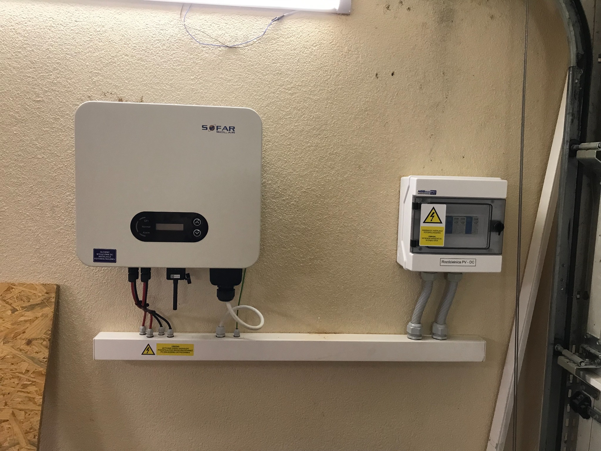 Biały falownik SOFAR i rozdzielnica PV-DC zamontowane na beżowej ścianie w garażu, widoczne przewody i ostrzeżenia o napięciu.