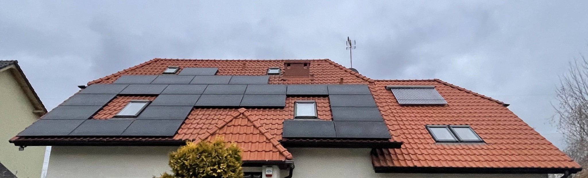 Czerwony dach domu z zamontowanymi panelami fotowoltaicznymi i kolektorami słonecznymi, widoczne okna dachowe, pochmurne niebo w tle.
