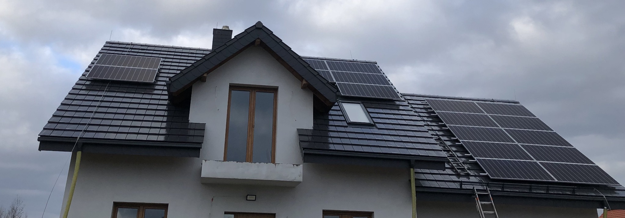 Dom z zainstalowanymi panelami fotowoltaicznymi na dachu pokrytym ciemną dachówką, widoczna drabina oparta o dach.