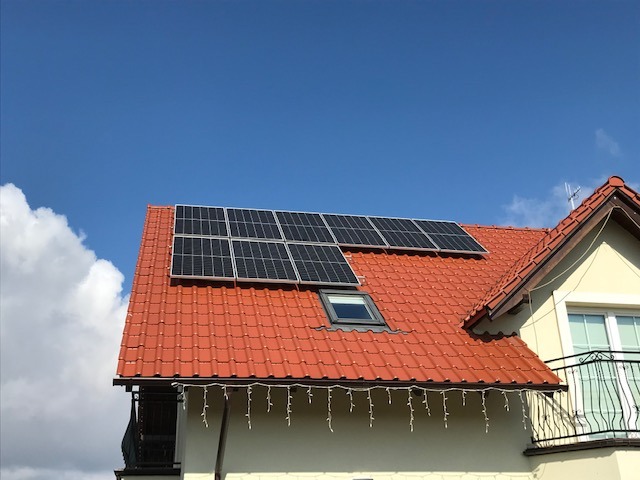 Panele słoneczne na dachu domu z czerwoną dachówką i oknem dachowym, błękitne niebo w tle.