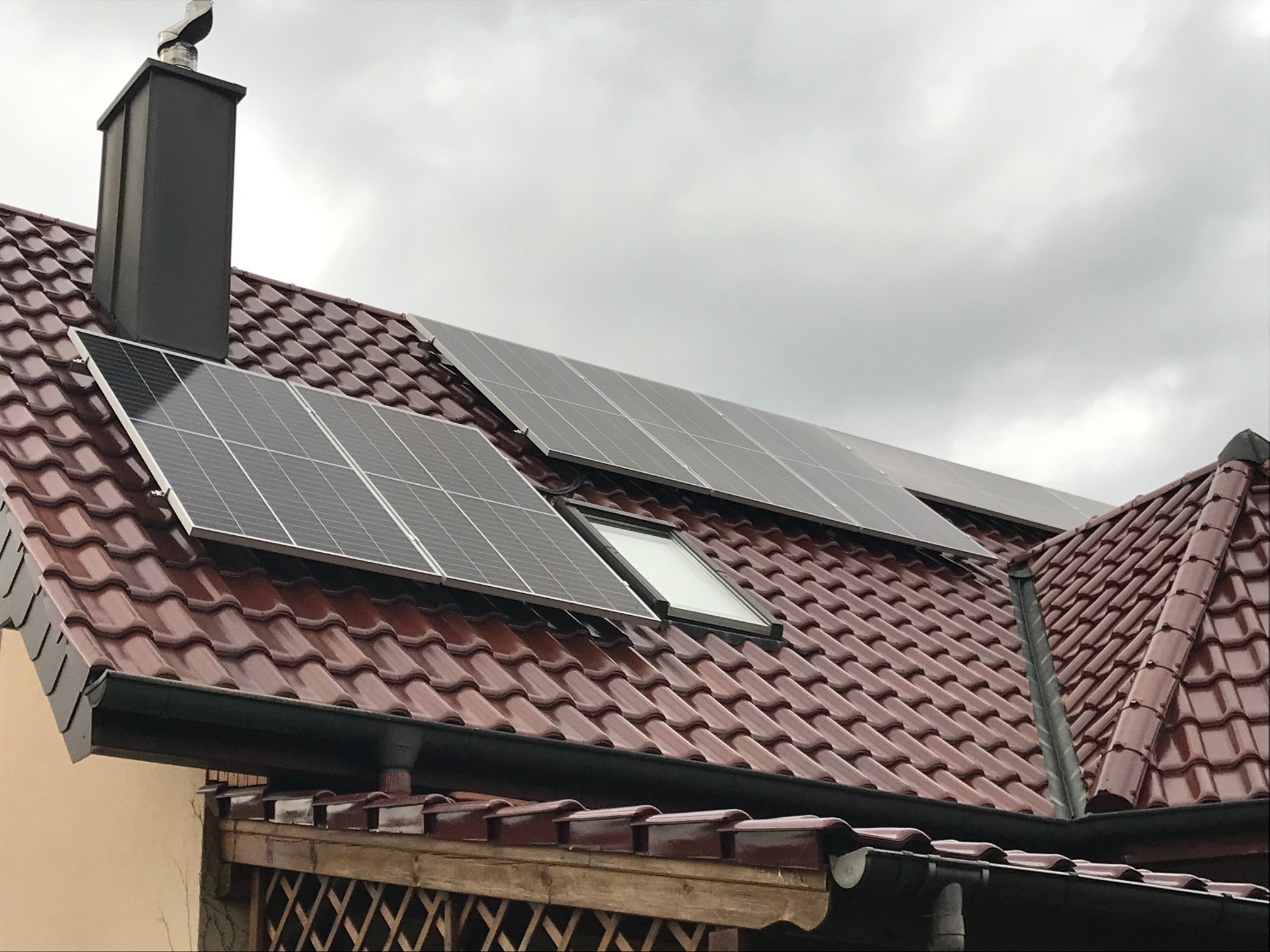Panele słoneczne na dachu domu z brązową dachówką, widoczne okno dachowe i komin na tle pochmurnego nieba.