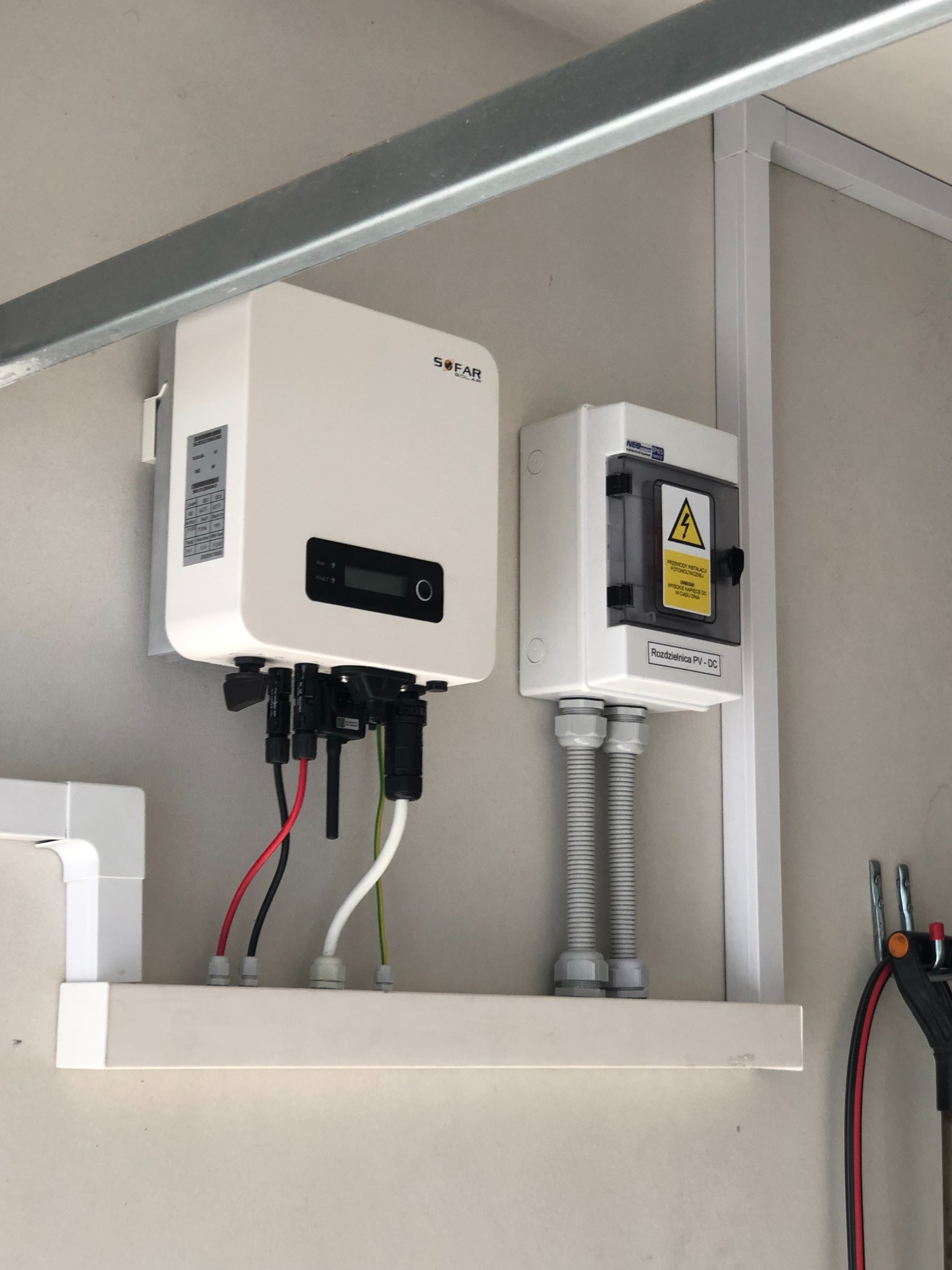 Biały inwerter marki SOFAR z podłączonymi kablami i biała rozdzielnica PV-DC z żółtym znakiem ostrzegawczym wysokiego napięcia, zamontowane na ścianie wewnątrz budynku.