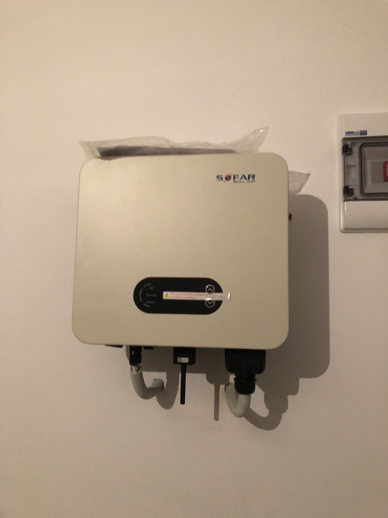 Biały falownik SOFAR Solar zamontowany na jasnej ścianie, z widocznymi przewodami i częściowo zdjętą folią ochronną, obok skrzynki elektrycznej.