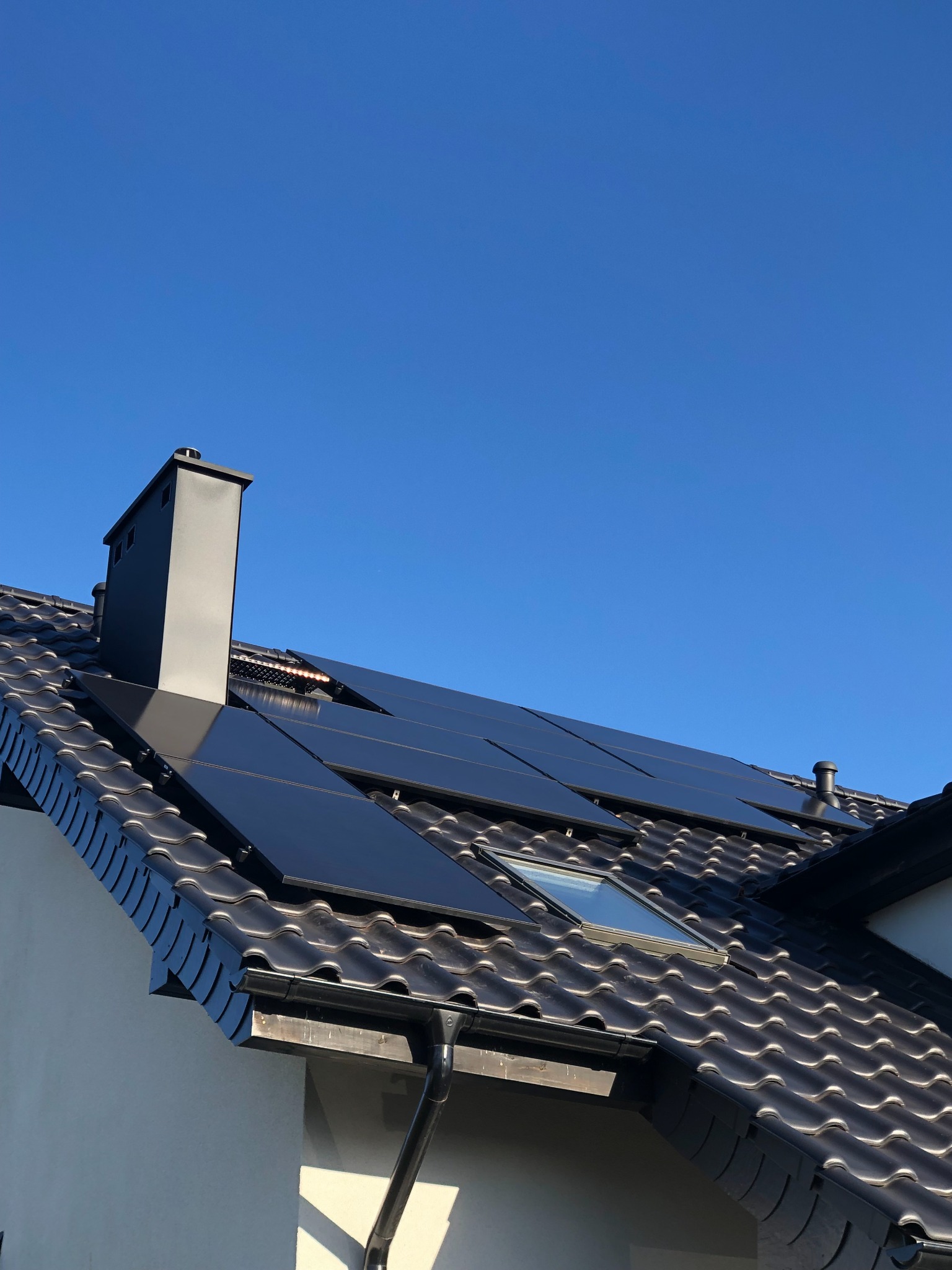 Czarne panele fotowoltaiczne na dachu z dachówki ceramicznej, widoczne okno dachowe i komin na tle błękitnego nieba.