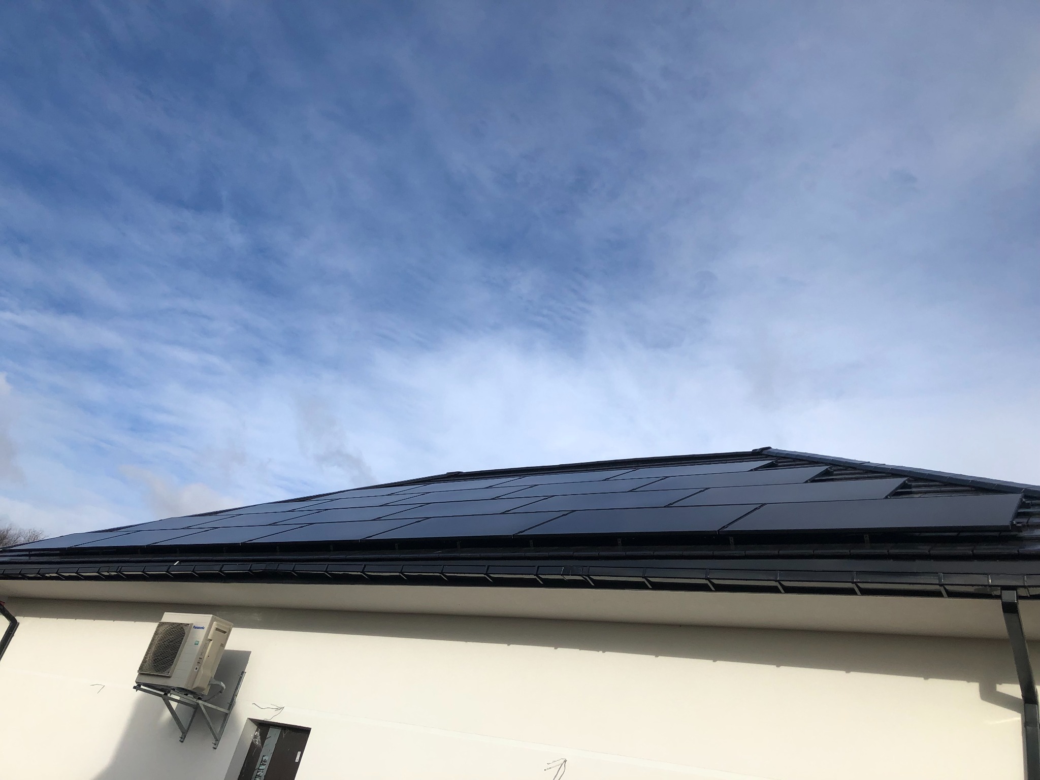 Panele słoneczne na dachu domu jednorodzinnego, widoczny klimatyzator na ścianie budynku i błękitne niebo z chmurami w tle.