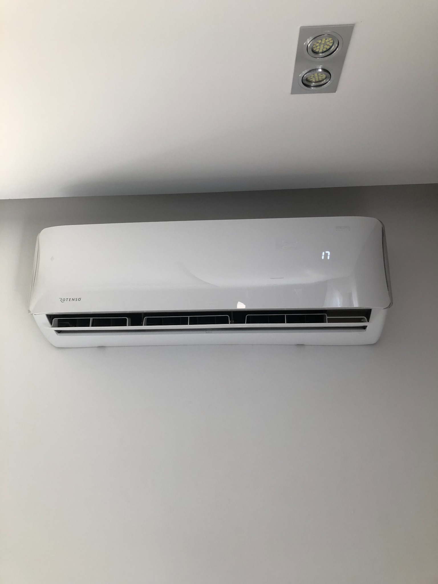 Biała jednostka wewnętrzna klimatyzatora marki Rotenso zamontowana na szarej ścianie, wyświetlająca temperaturę 17 stopni, oświetlona przez sufitowe reflektory.