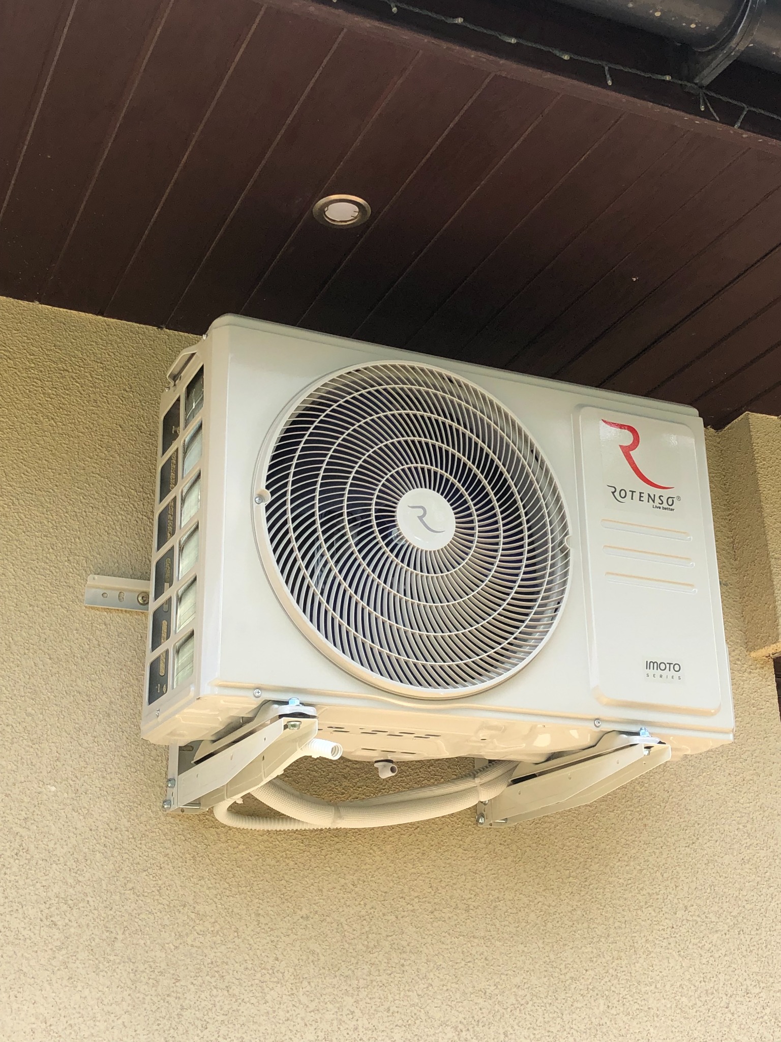 Zewnętrzna jednostka klimatyzacji Rotenso Imoto Series zamontowana na ścianie budynku o beżowej, chropowatej fakturze, z widocznymi przewodami i elementami montażowymi.