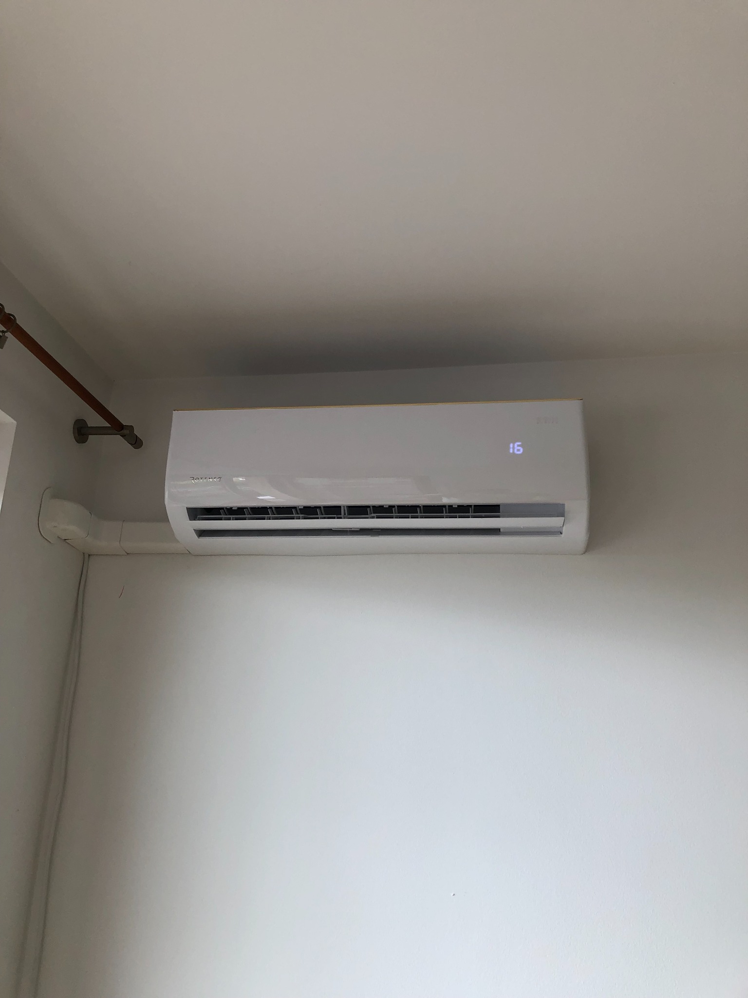 Biała jednostka klimatyzacyjna zamontowana na białej ścianie, z widocznym wyświetlaczem ustawionej temperatury 16 stopni Celsjusza i rurami odprowadzającymi skropliny.