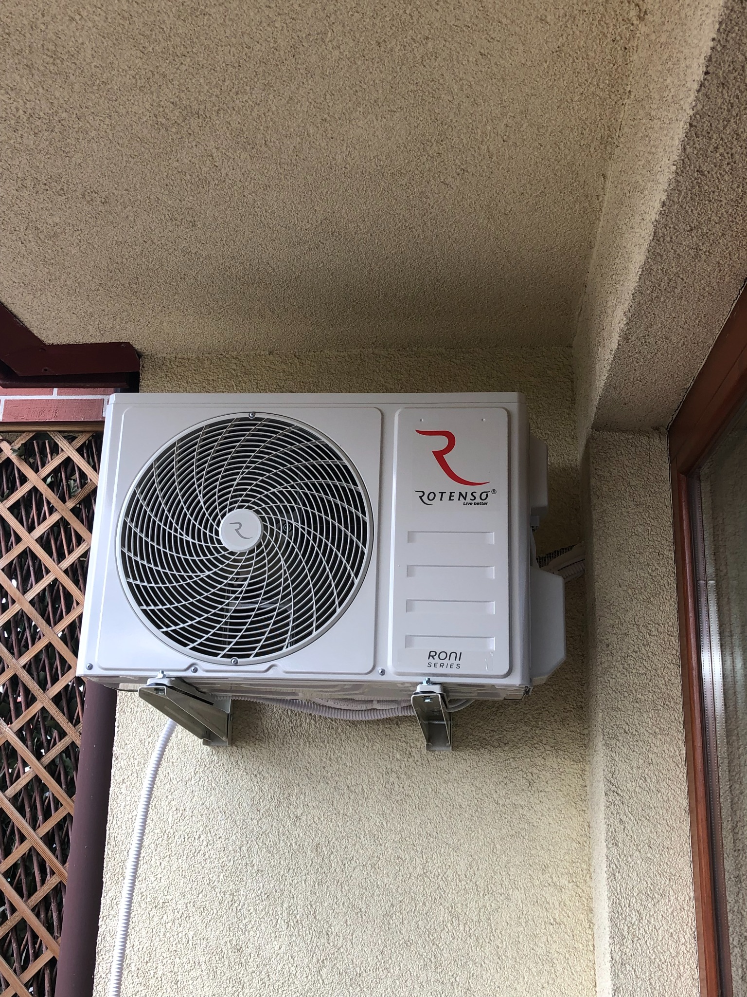 Biała jednostka zewnętrzna klimatyzatora marki Rotenso, model RONI, zamontowana na teksturowanej elewacji budynku, widoczna z boku okna i drewnianej kraty.