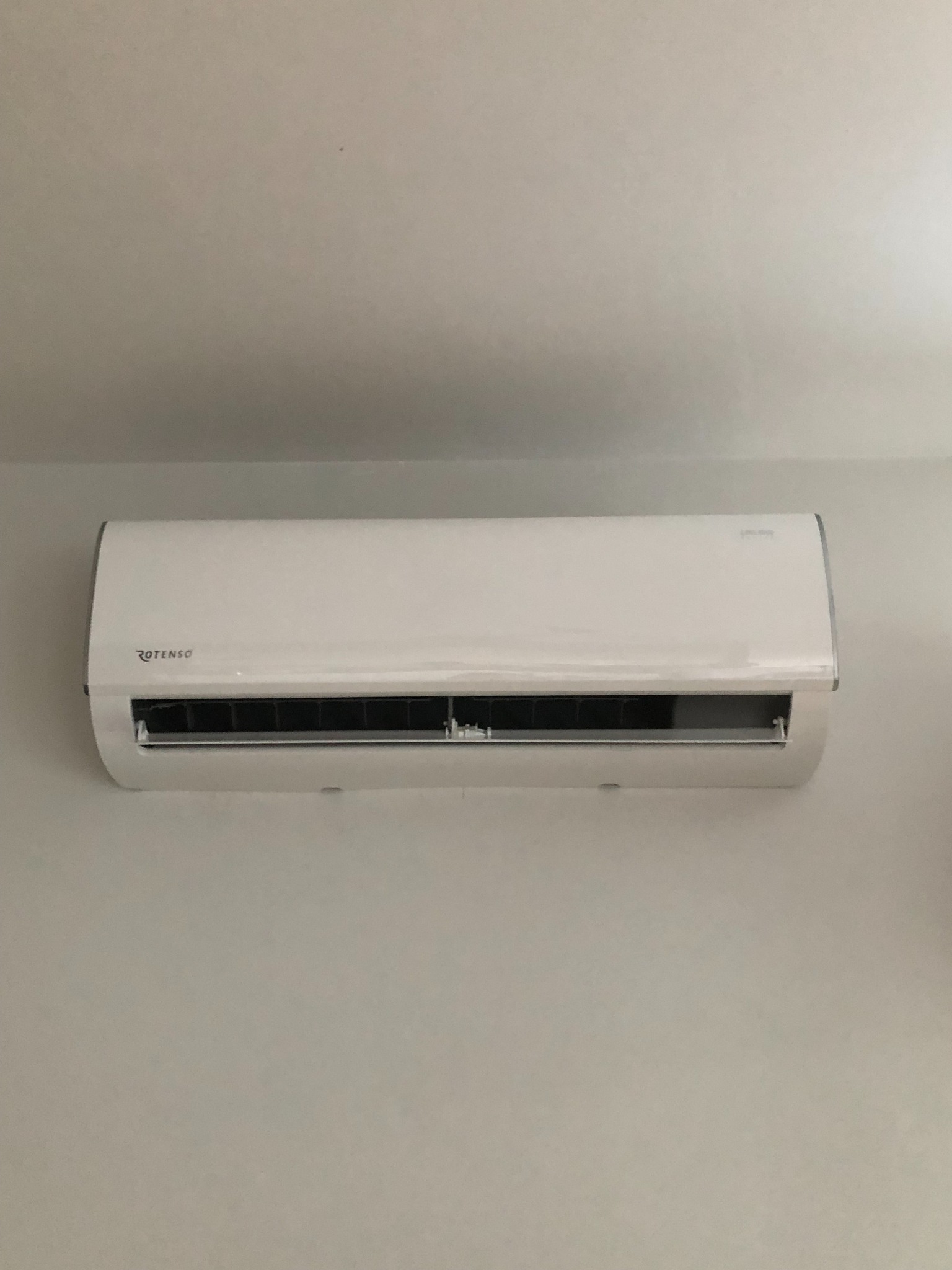 Biała jednostka wewnętrzna klimatyzatora marki Rotenso zamontowana na jasnej ścianie, widoczna z przodu, z delikatnym światłem padającym z góry.
