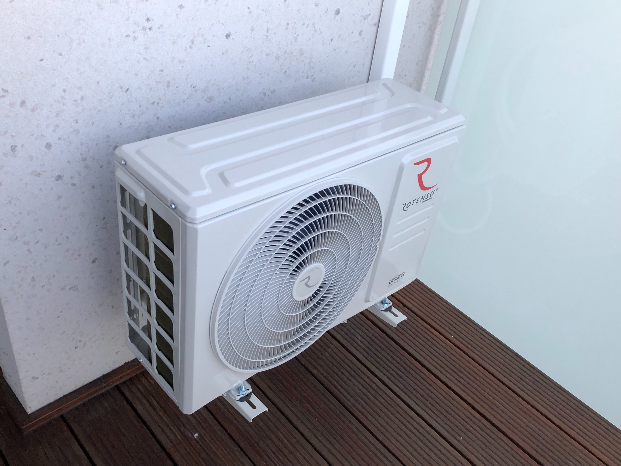 Zewnętrzna jednostka klimatyzacji Rotenso Ukura Series zamontowana na drewnianym tarasie przy ścianie z jasnego kamienia, widok z góry.