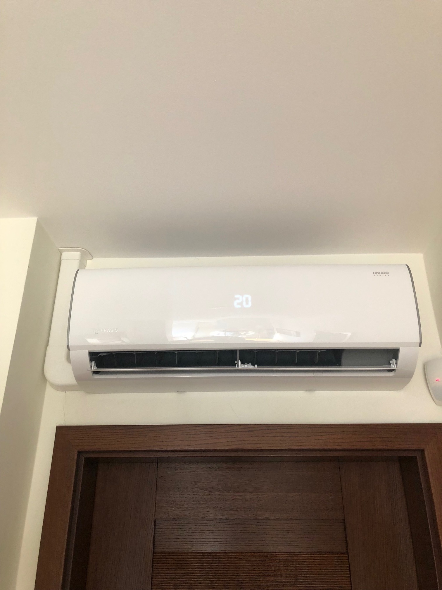 Biały klimatyzator Ukura Master zamontowany nad ciemnobrązowymi drzwiami, wyświetlający temperaturę 20 stopni Celsjusza.