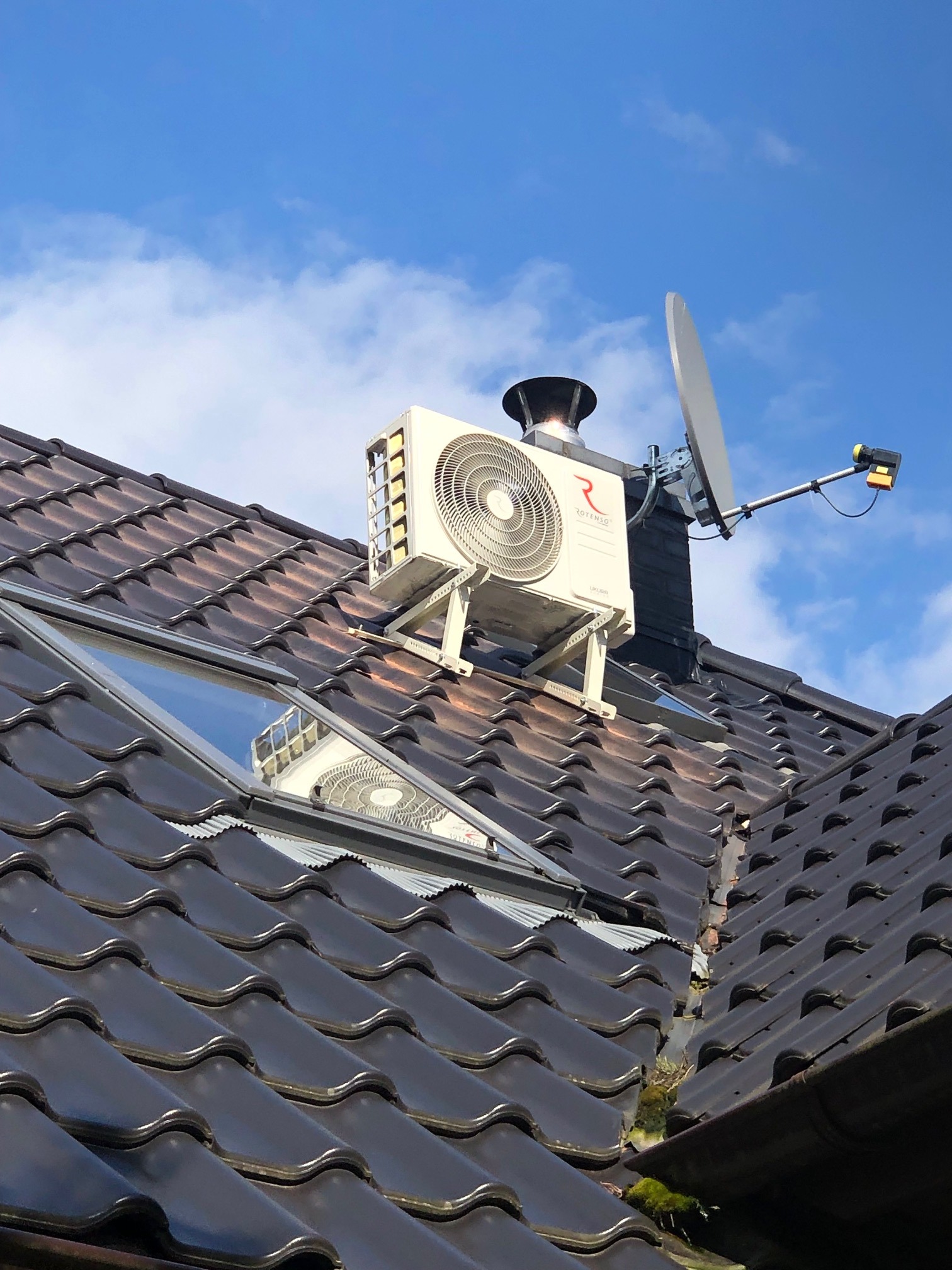 Biała jednostka zewnętrzna klimatyzatora marki Rotenso zamontowana na dachu z ciemnej dachówki obok komina i anteny satelitarnej, widok na niebieskie niebo z chmurami w tle.