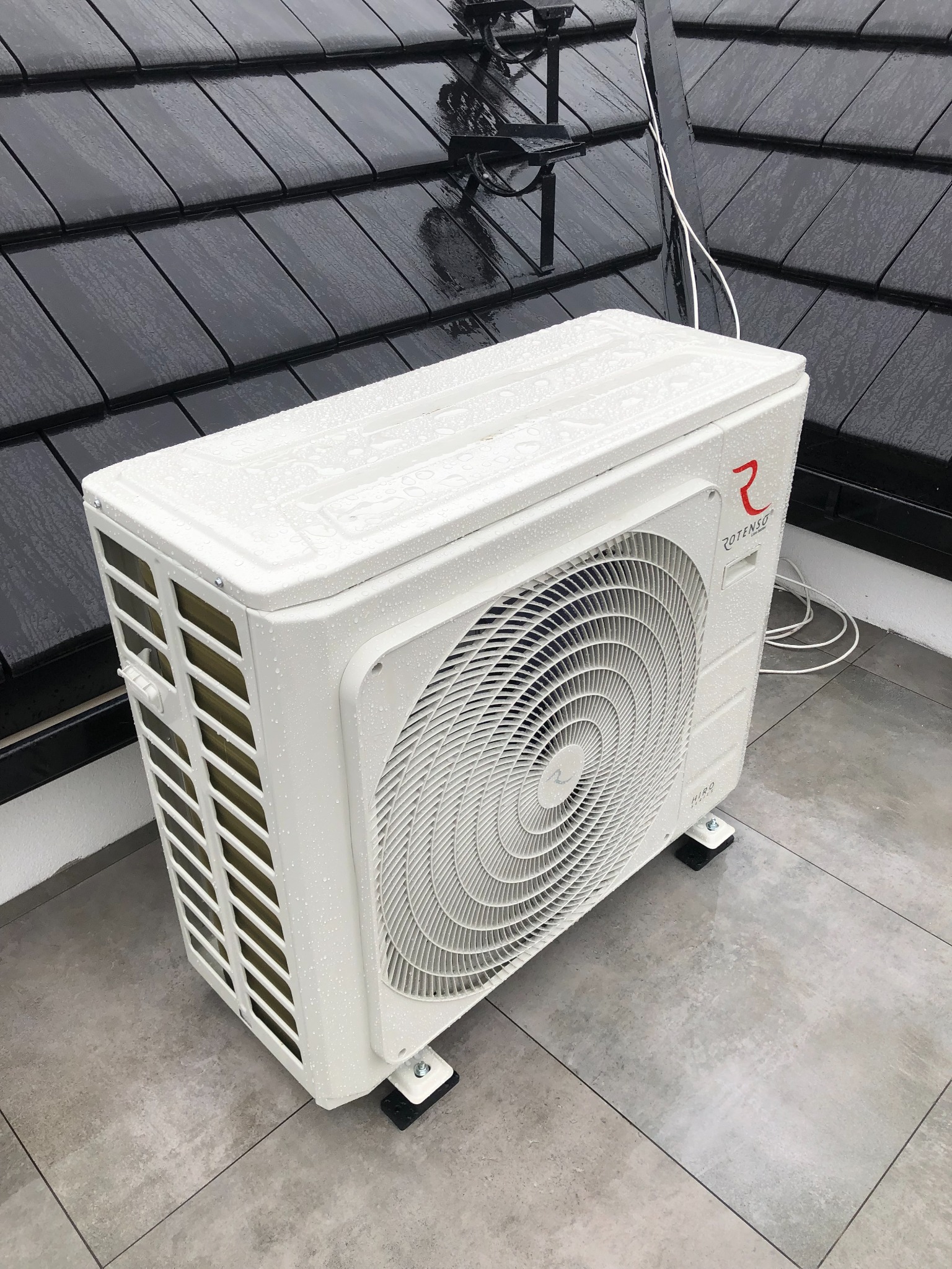 Zewnętrzna jednostka klimatyzacji Rotenso HIRO, umieszczona na szarych płytkach tarasowych, z czarnym dachem w tle, pokryta kroplami deszczu.
