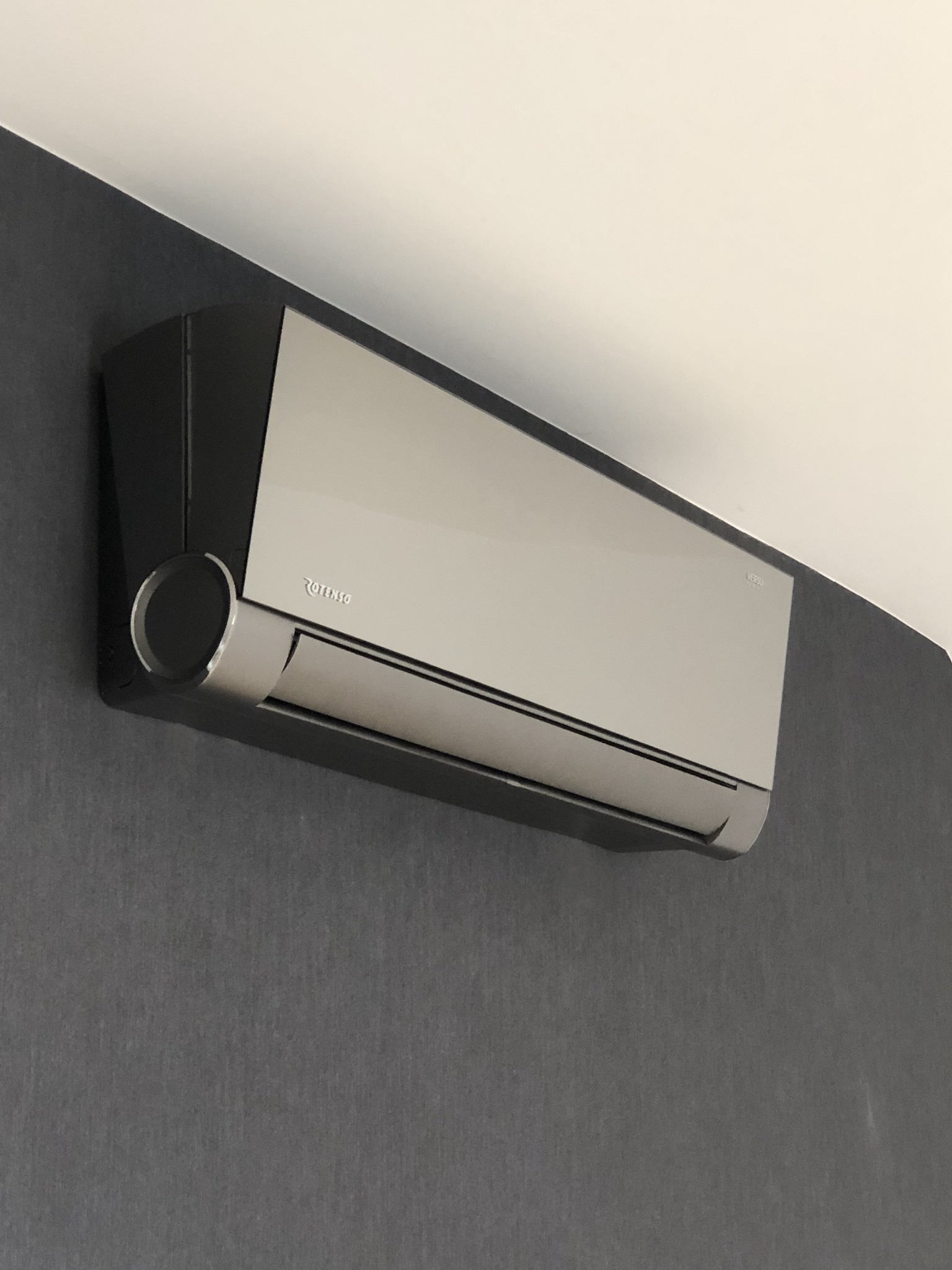 Szaro-czarna jednostka wewnętrzna klimatyzatora marki Rotenso zamontowana na ciemnoszarej ścianie, widok z dołu.