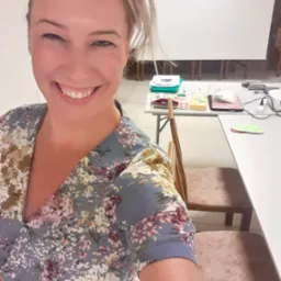 Uśmiechnięta kobieta w kwiecistej sukience robi selfie na tle sali szkoleniowej z białym ekranem i stołami z materiałami edukacyjnymi.