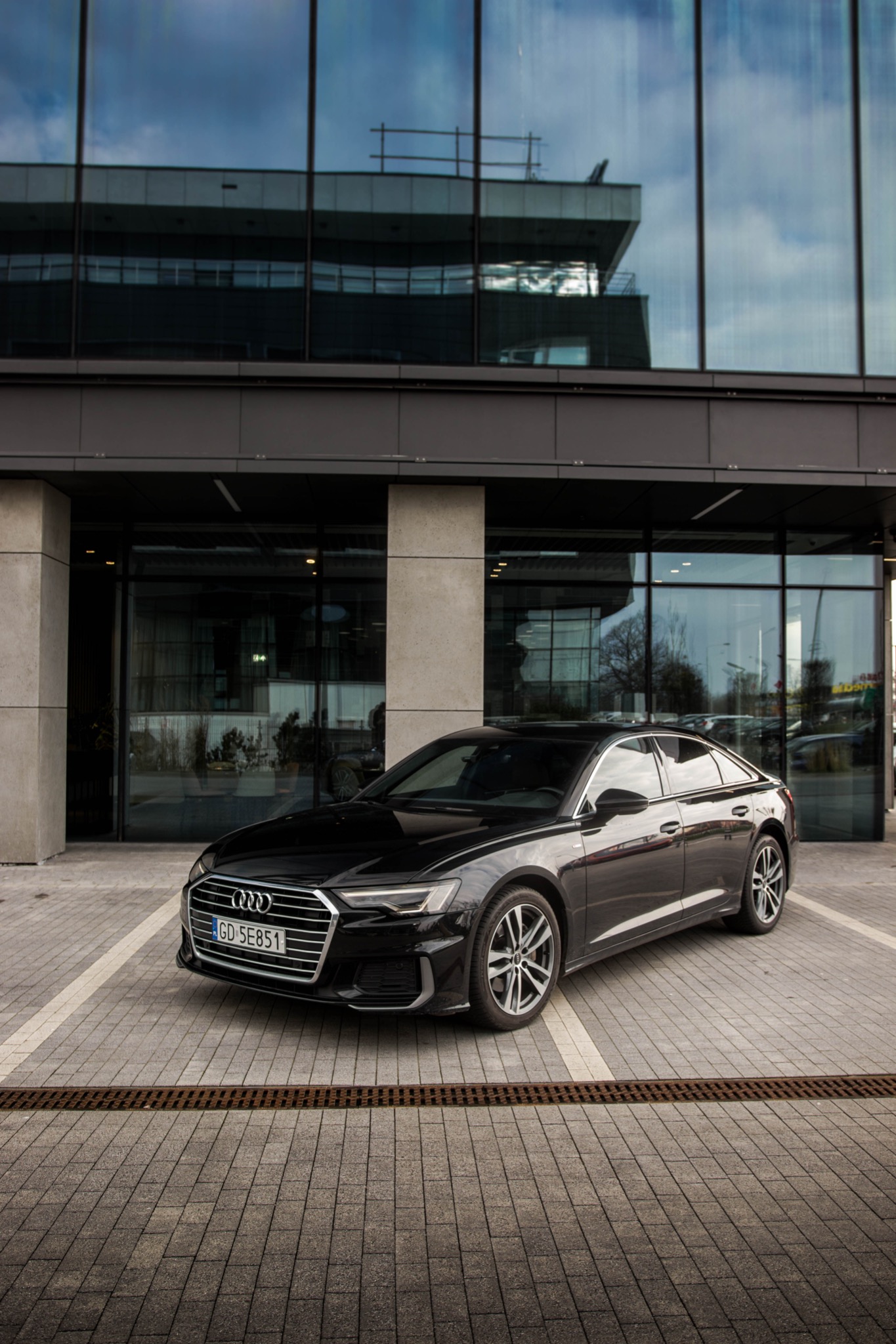 Czarna limuzyna Audi zaparkowana przed nowoczesnym budynkiem biurowym z szklaną fasadą odbijającą niebo, widoczna tablica rejestracyjna GD 5E851.