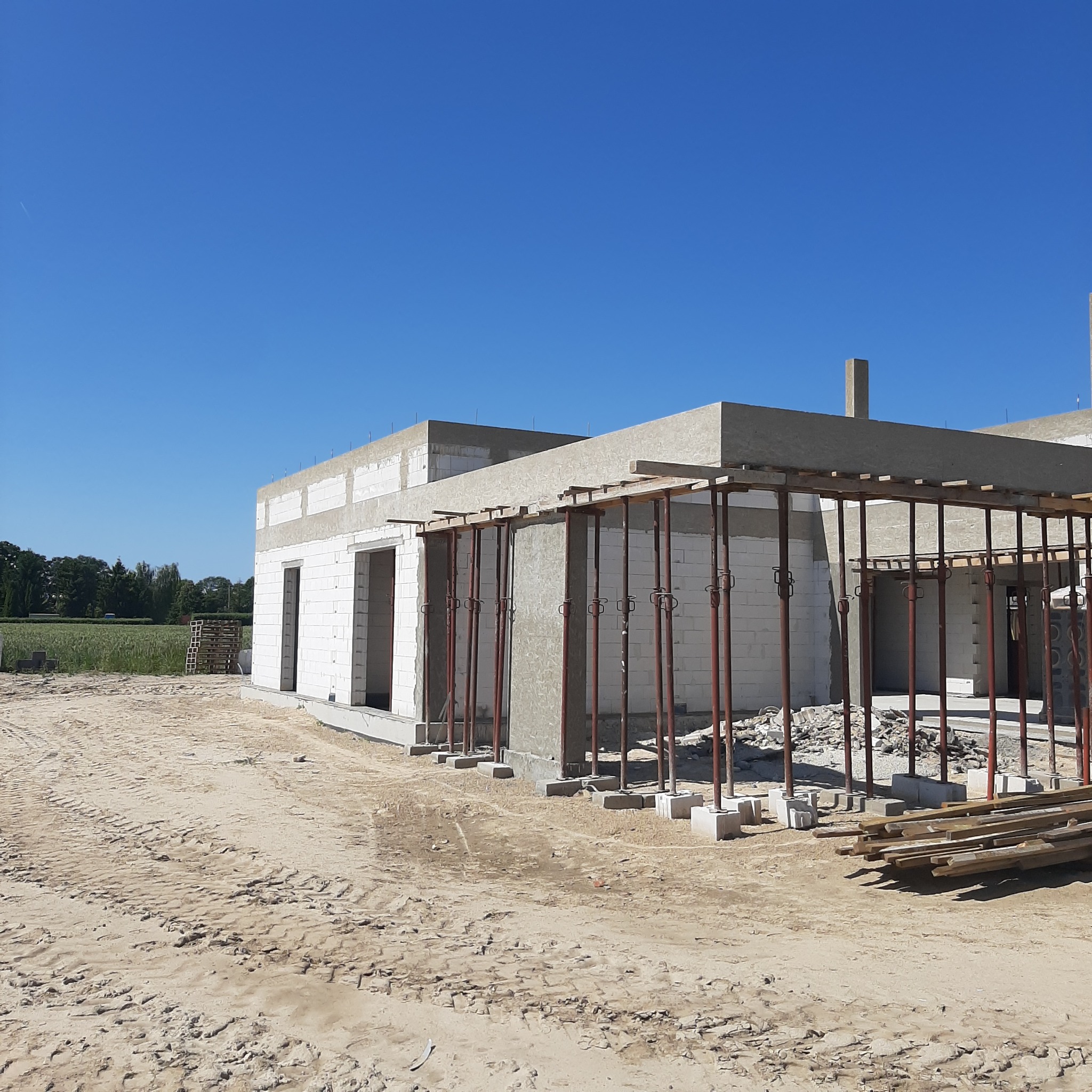 Wznoszenie ścian domu z betonu komórkowego, widoczne podpory stropu. W tle zielone pola i błękitne niebo w słoneczny dzień. Etap budowy.