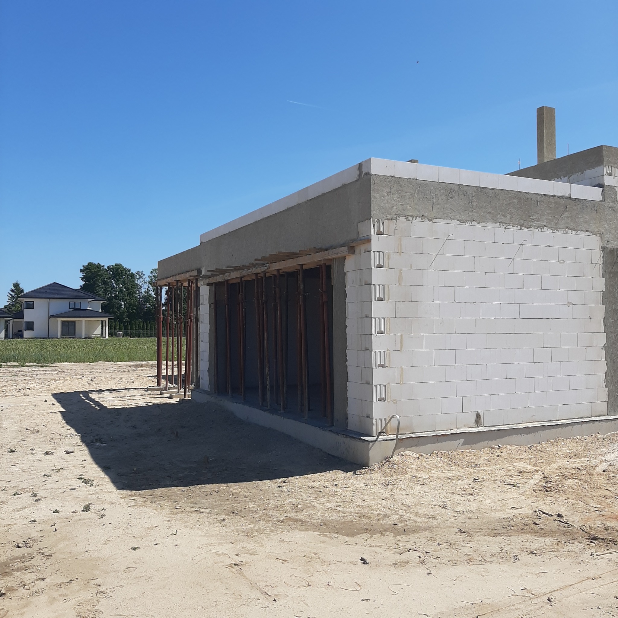 Budowa domu z białych bloczków, widoczne podpory szalunkowe. W tle dom jednorodzinny i błękitne niebo. Stan surowy otwarty.