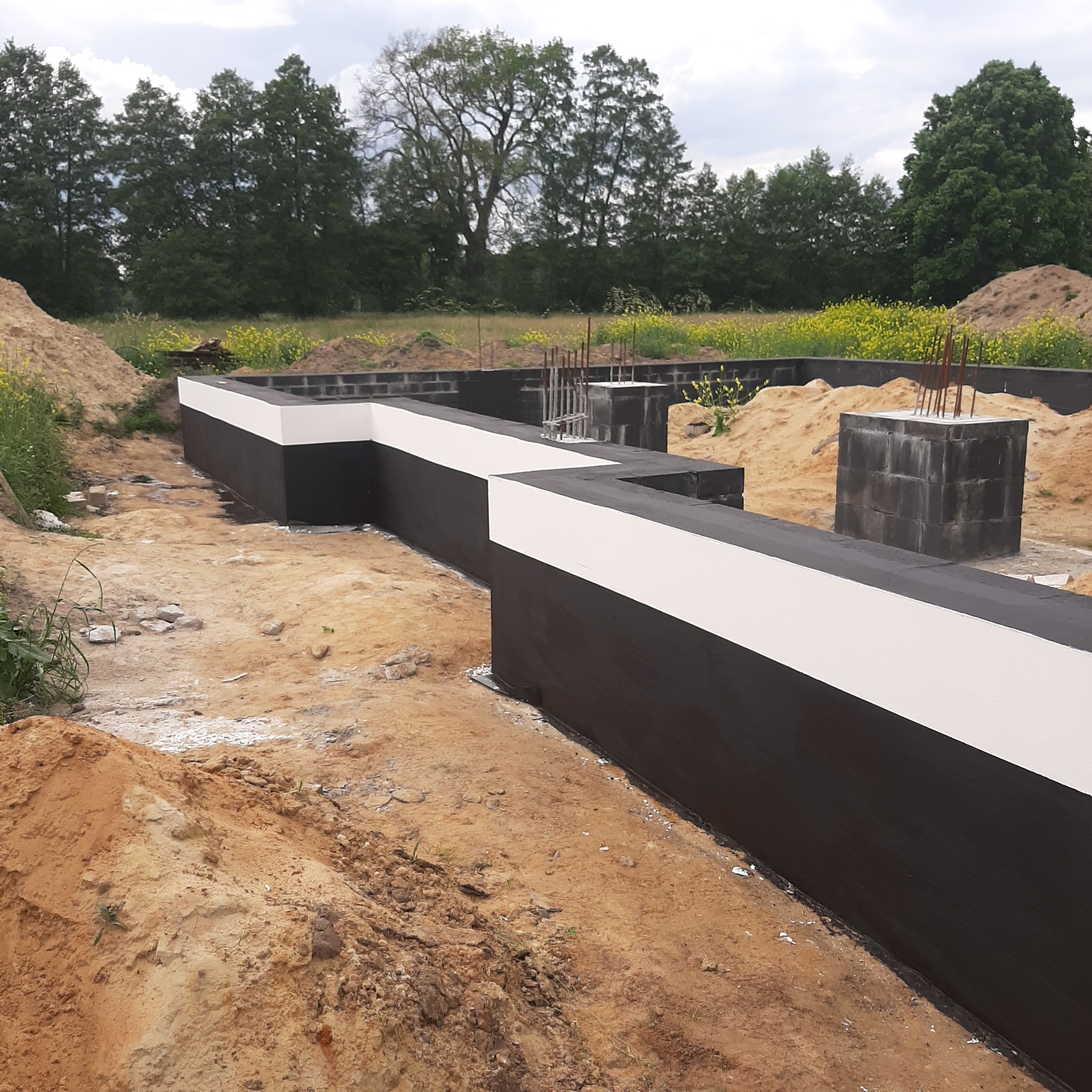 Zabezpieczone, zaizolowane fundamenty domu z bloczków betonowych, częściowo zasypane ziemią. Widoczne zbrojenia i roślinność w tle.