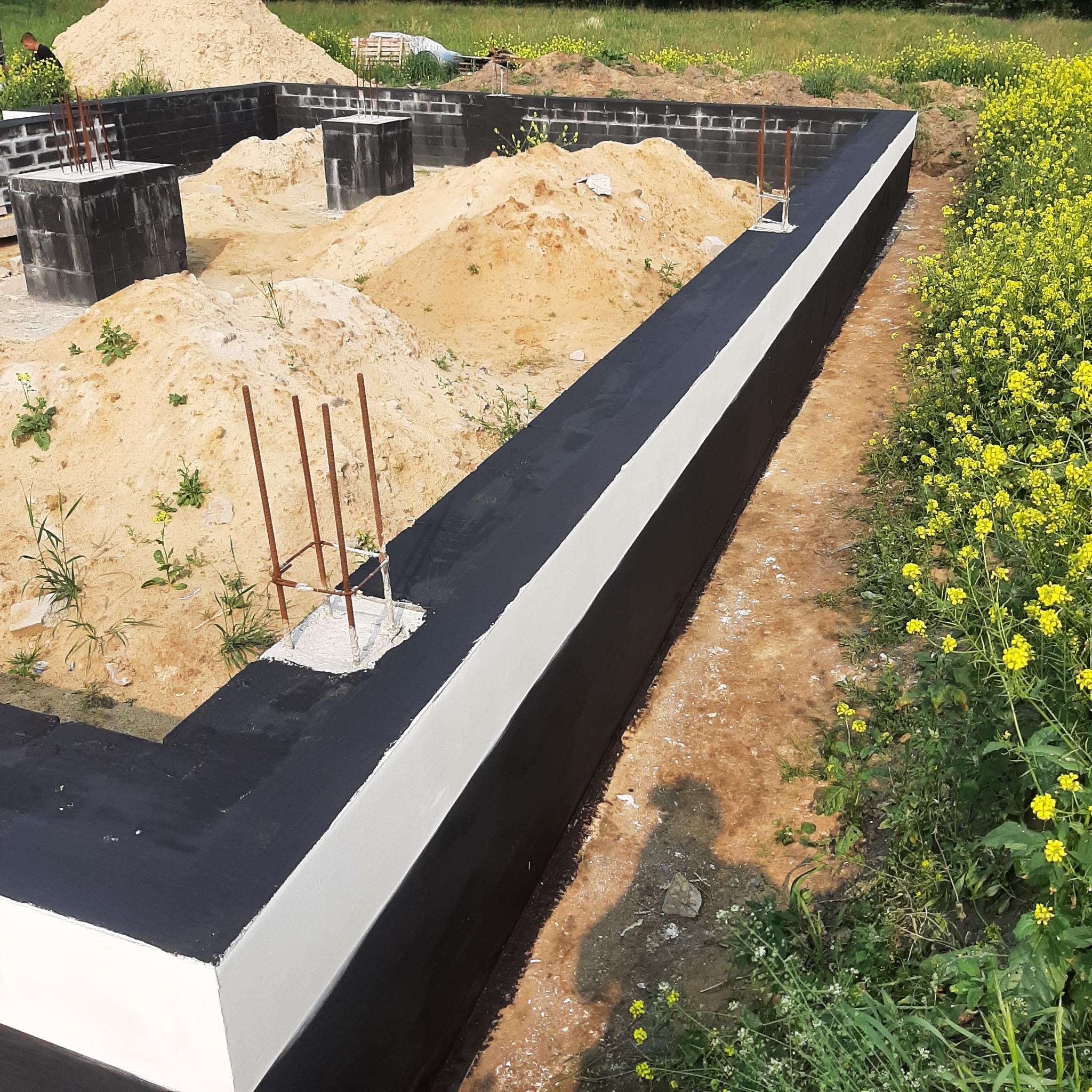 Zabezpieczone fundamenty domu z widocznym zbrojeniem, przygotowane do dalszych prac budowlanych. Na pierwszym planie izolacja przeciwwilgociowa.