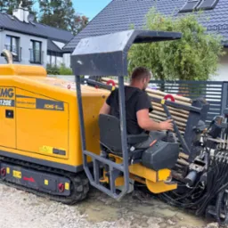 Żółta maszyna XCMG XZ120E do przewiertów sterowanych z operatorem na tle domu i ogrodzenia. Widoczne rury wiertnicze i przewody hydrauliczne.