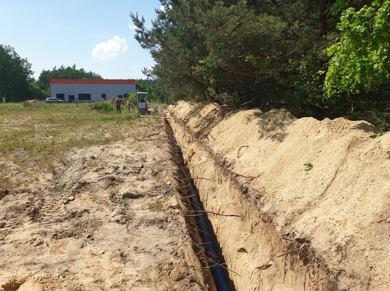 Długi wykop w piaszczystej ziemi, w którym ułożona jest rura. Na dalszym planie widoczny mały traktor i dwie osoby. W tle las i budynek.