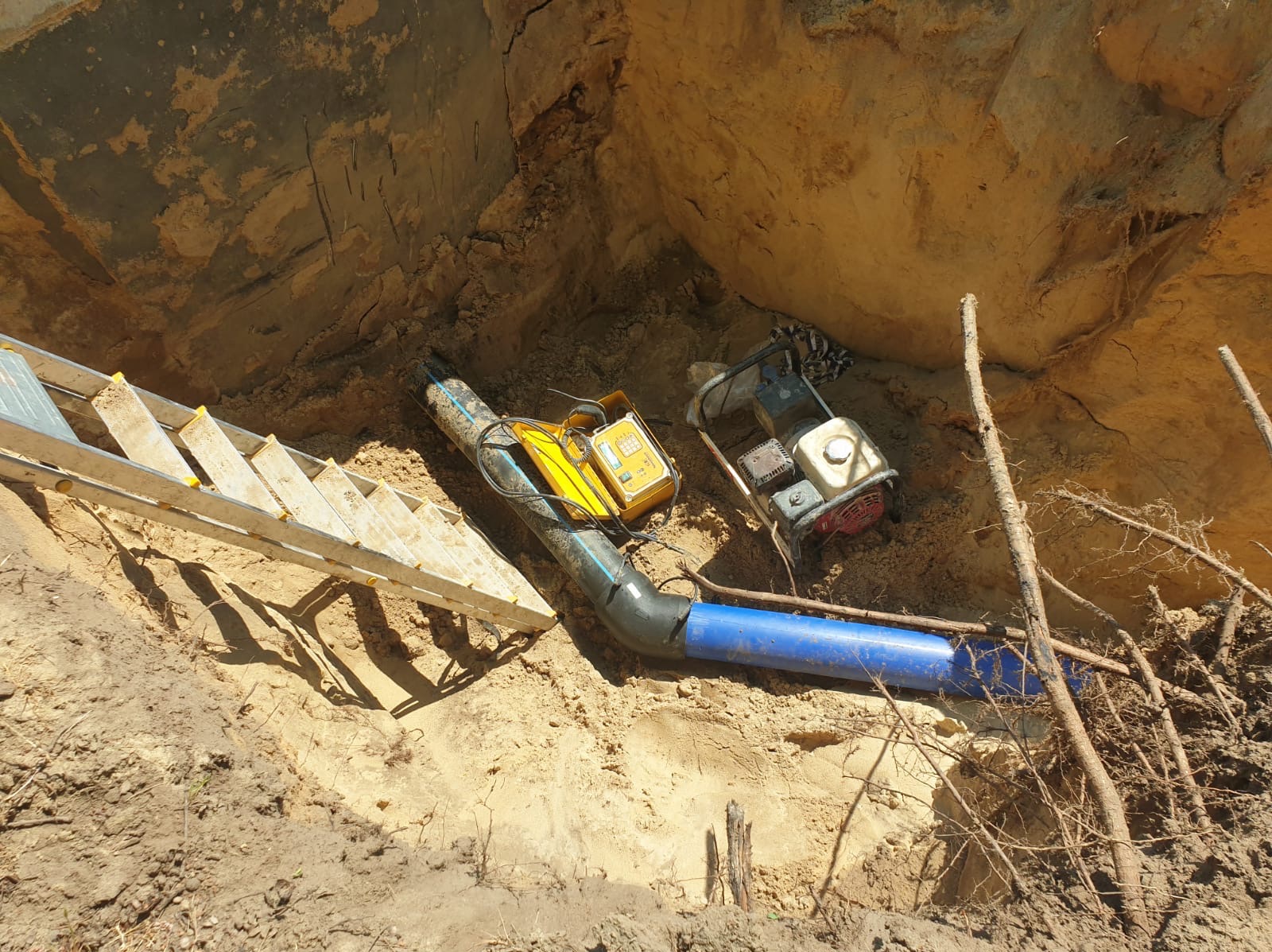 Wykop z ułożoną rurą wodociągową z niebieskiego tworzywa, ubijarka wibracyjna i detektor przewodów w wykopie, drabina przystawna.