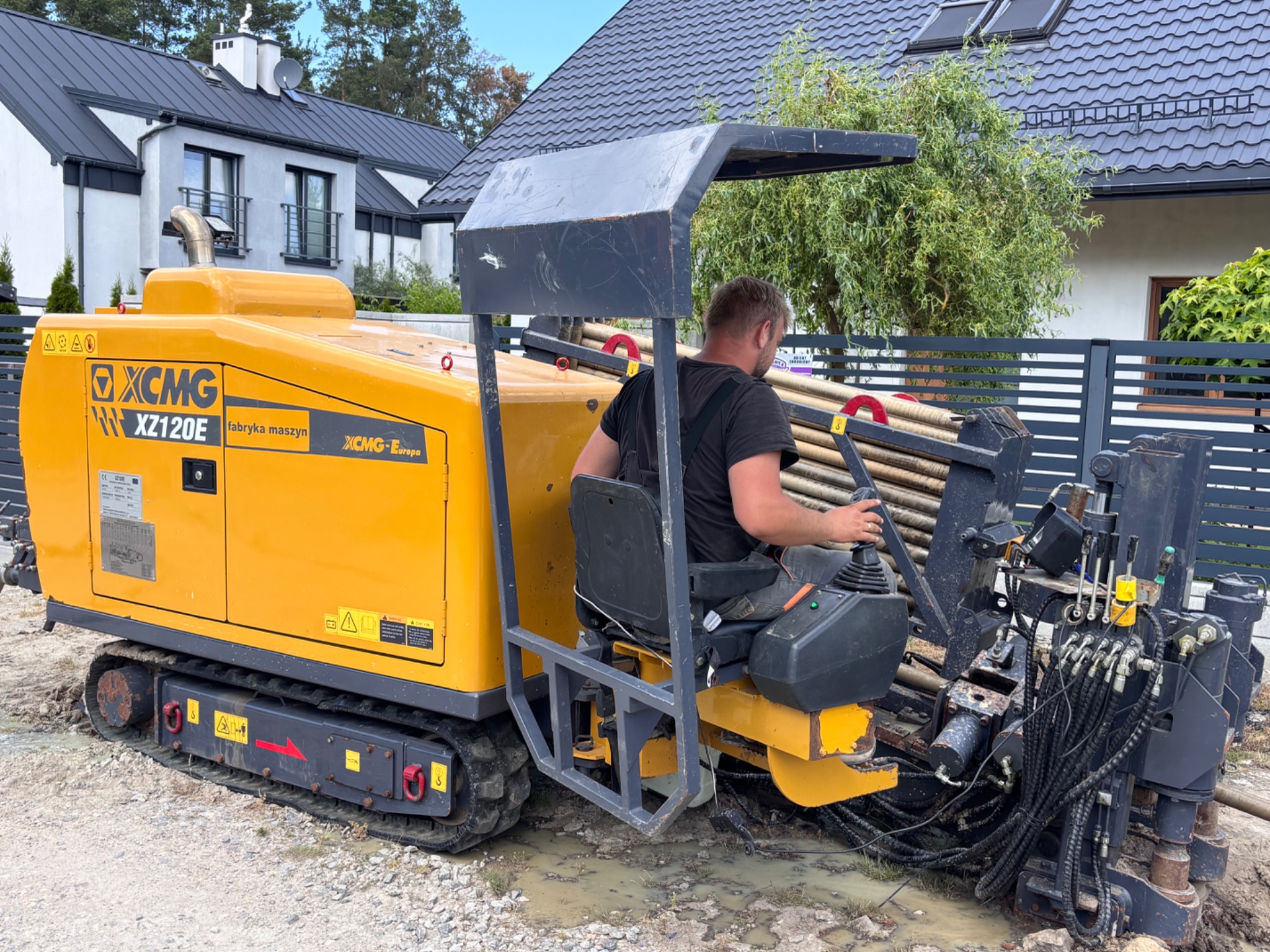 Żółta maszyna XCMG XZ120E do przewiertów sterowanych z operatorem na tle domu i ogrodzenia. Widoczne rury wiertnicze i przewody hydrauliczne.