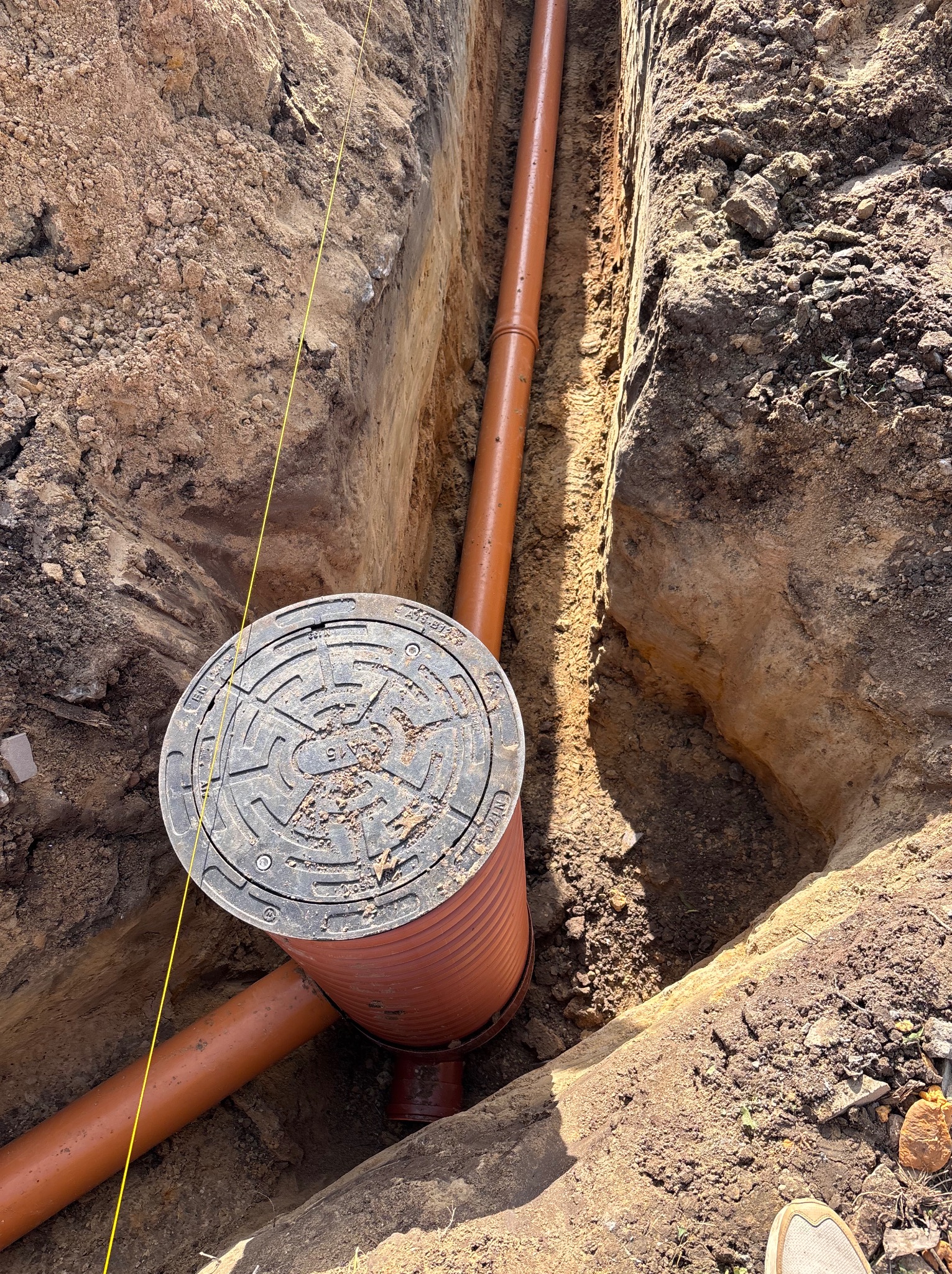 Pomarańczowa rura kanalizacyjna w wykopie ziemnym, z widocznym włazem studzienki. Żółta linka wyznacza poziom. Ujęcie z góry, podkreślające głębokość wykopu.