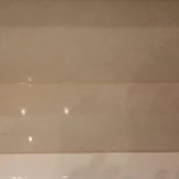 Beżowe, poziome płytki ceramiczne na ścianie łazienki, widoczne spoiny i fragment białej wanny w dolnej części kadru, oświetlenie halogenowe odbijające się od powierzchni płytek.