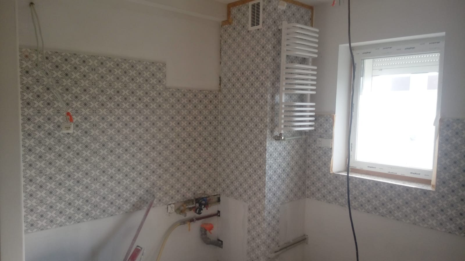 Wnętrze łazienki w trakcie remontu, z częściowo położonymi płytkami w geometryczny wzór, grzejnikiem drabinkowym na ścianie, widocznymi instalacjami hydraulicznymi i oknem z roletą.