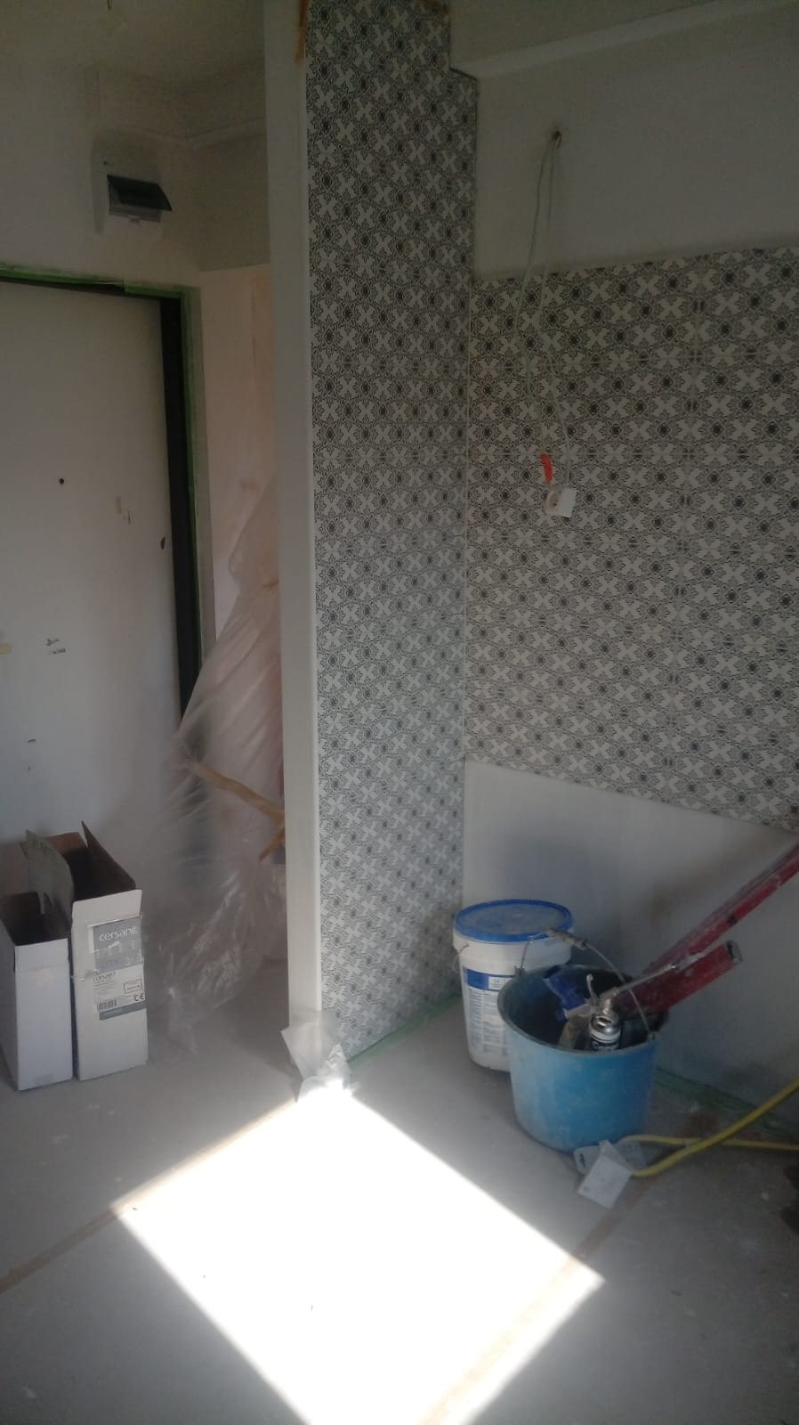 Fragment łazienki w trakcie prac wykończeniowych: ułożone płytki z wzorem na ścianie, wiadro z narzędziami, kartony z ceramiką Cersanit, zabezpieczona folią futryna drzwi.