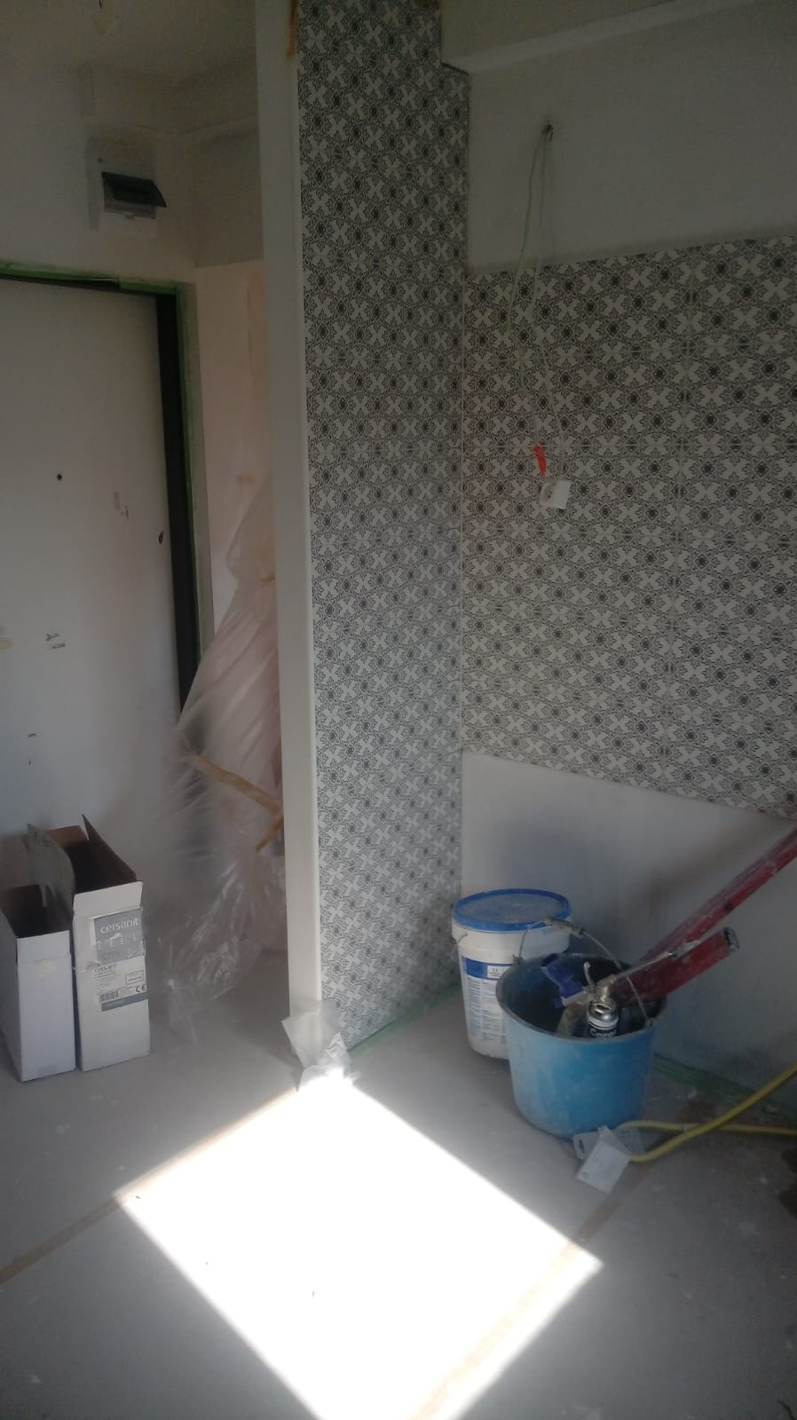 Fragment łazienki w trakcie prac z ułożoną częścią dekoracyjnej mozaiki na ścianie, widoczne wiadro z zaprawą, pojemnik z narzędziami, kartony po płytkach Cersanit oraz zabezpieczona folią futryna...