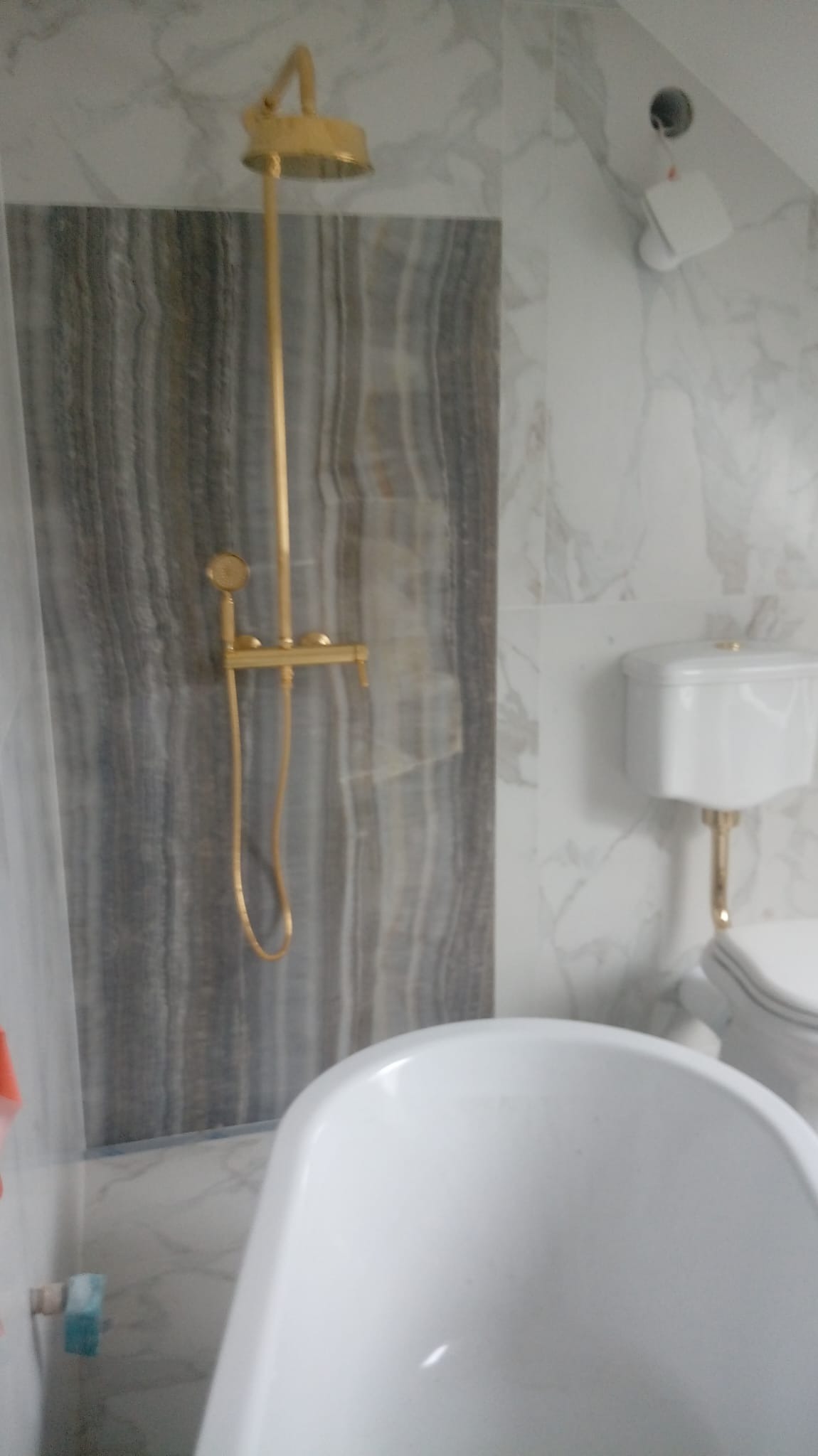 Nowoczesna łazienka z białą wanną, toaletą z elementami w kolorze złotym i deszczownicą w kolorze złotym na tle płytek imitujących marmur.