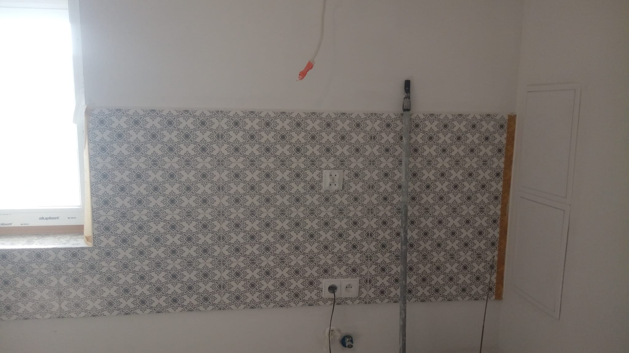 Fragment ściany z częściowo położoną dekoracyjną glazurą w odcieniach szarości i bieli, widoczne gniazdka elektryczne, rurka instalacyjna i okno.