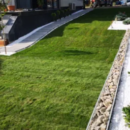 Starannie zaprojektowany ogród z równo przystrzyżonym trawnikiem, białymi kamieniami dekoracyjnymi, gabionami z kamienia i różnorodnymi nasadzeniami drzew i krzewów.