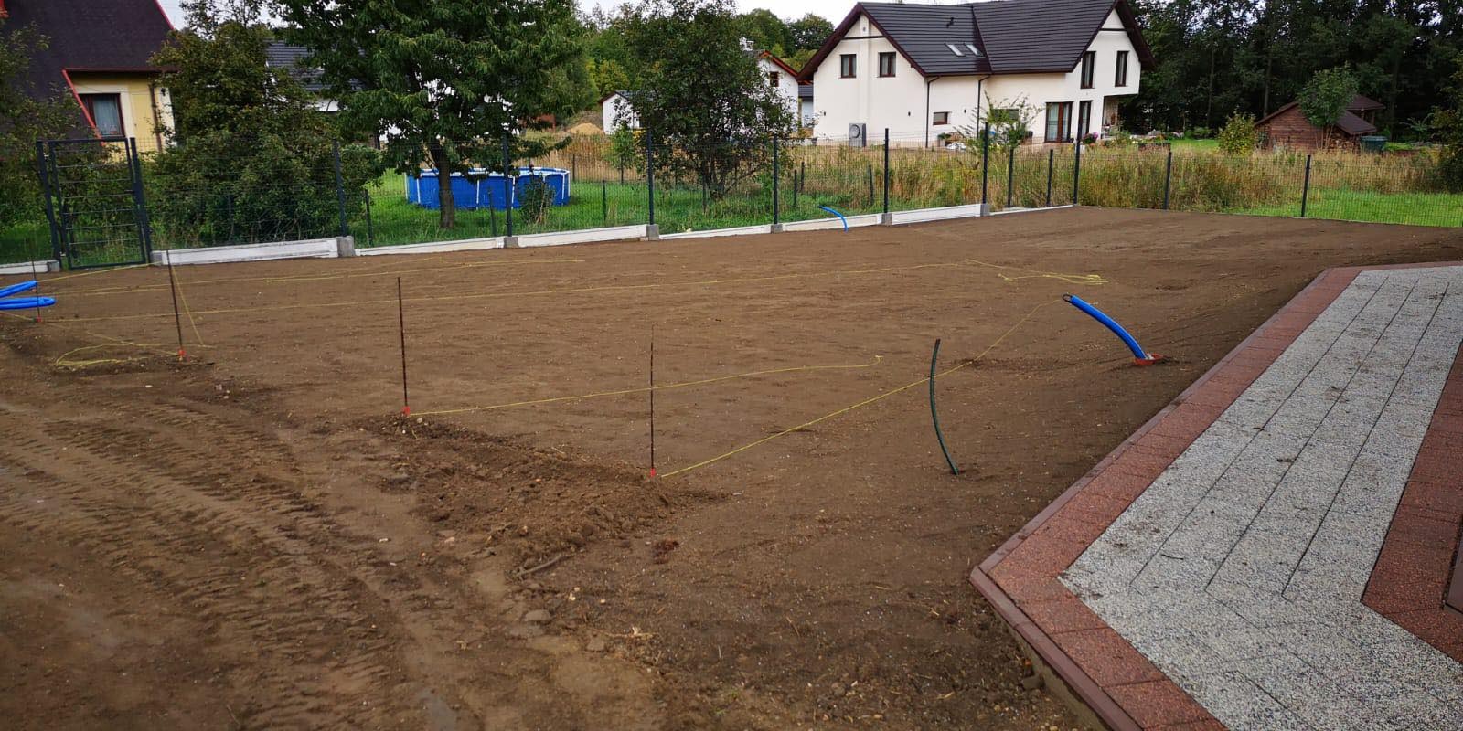Przygotowany teren pod przyszły ogród z wytyczonymi granicami, widoczne sznurki i paliki, w tle domy i drzewa.