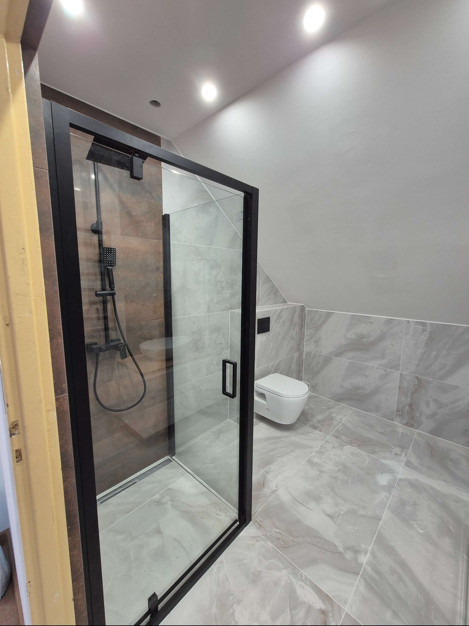 Wykończona łazienka z kabiną prysznicową typu walk-in z czarną ramą, marmurowe płytki na podłodze i ścianach, wisząca toaleta. Nowoczesny design.