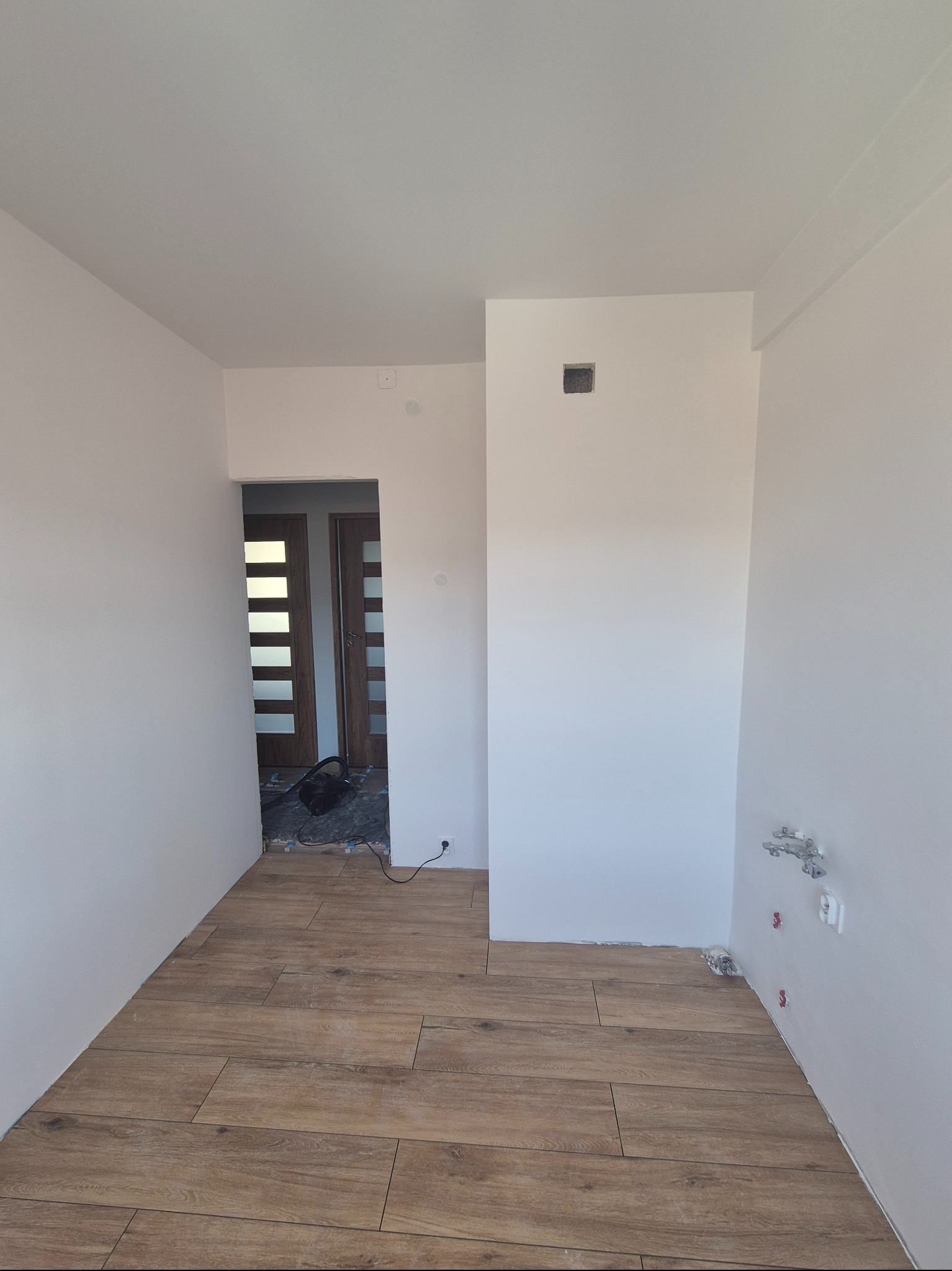 Jasne wnętrze po remoncie: drewnopodobna podłoga, świeżo pomalowane ściany, widoczne instalacje hydrauliczne i elektryczne. W tle otwarte drzwi do kolejnego pomieszczenia.