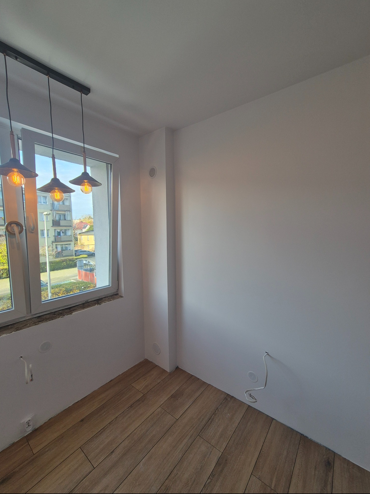 Jasne wnętrze po remoncie: drewnopodobna podłoga, białe ściany z gniazdkami i widok z okna z ozdobnymi lampami. Minimalistyczny design.