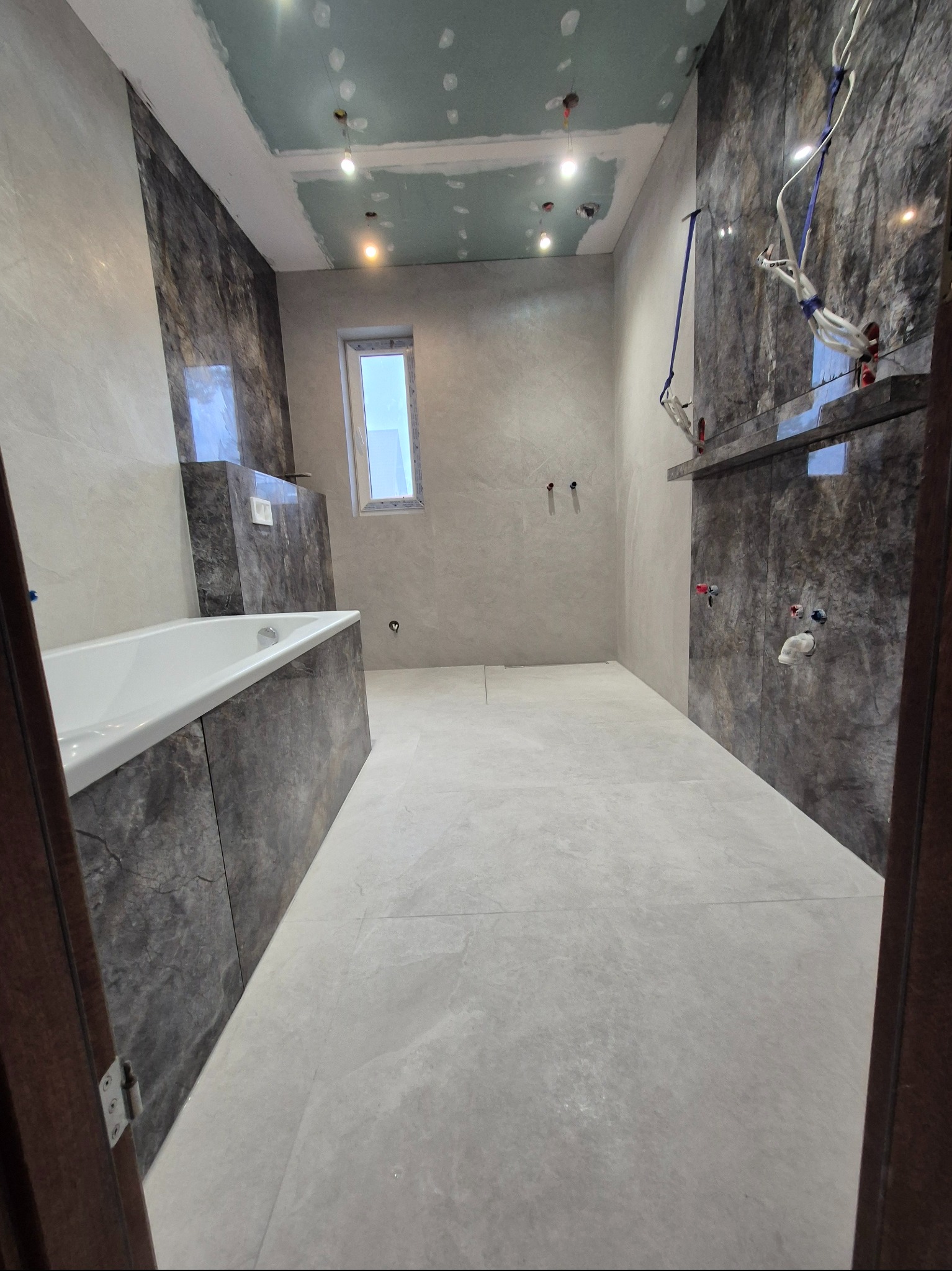 Wnętrze łazienki w trakcie wykańczania: położone płytki podłogowe i ścienne, częściowo zabudowana wanna, sufit z płyt gipsowo-kartonowych, widoczne instalacje.