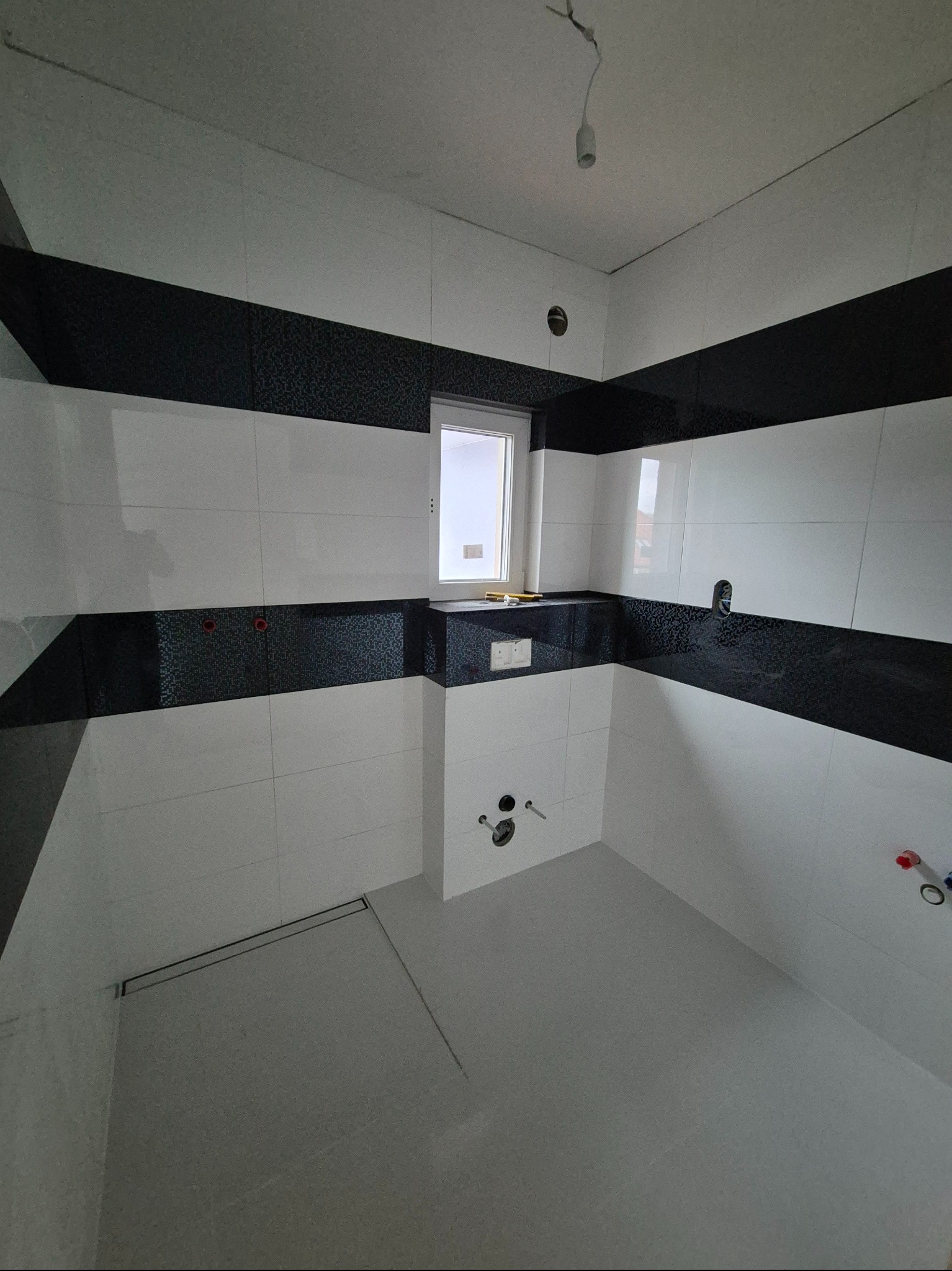 Wykończona łazienka z czarno-białymi pasami płytek, widoczny odpływ liniowy i instalacja pod armaturę. Minimalistyczny design z oknem.