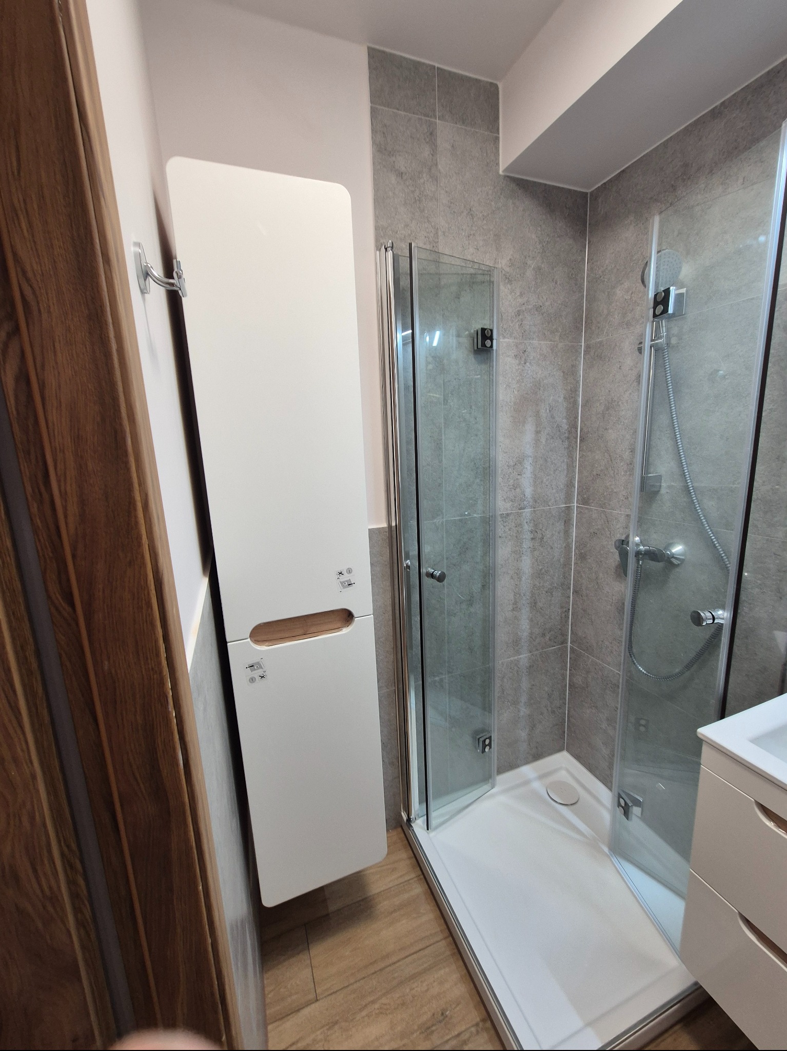 Nowoczesna łazienka z kabiną prysznicową, szarą glazurą i białymi meblami. Uwagę zwraca wysoka szafka łazienkowa z drewnianym akcentem i drewnopodobna podłoga.