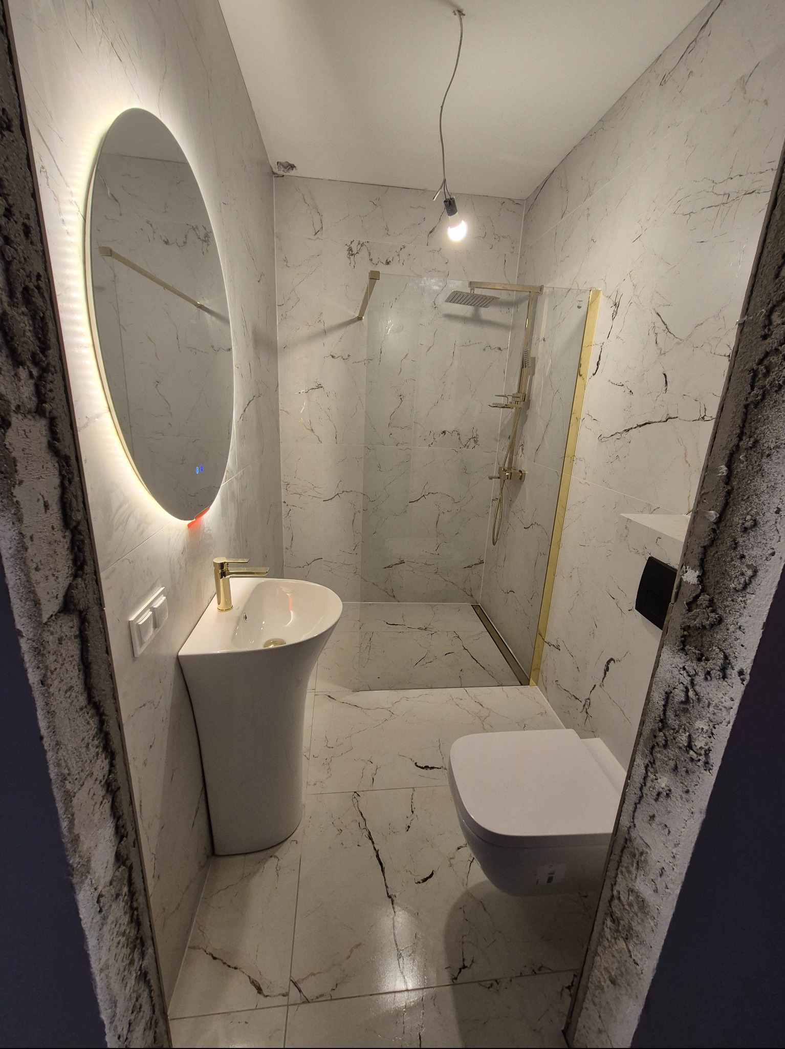 Nowoczesna łazienka z marmurowymi płytkami, złotymi akcentami i podświetlanym lustrem owalnym. Widok na kabinę prysznicową i wiszącą toaletę.