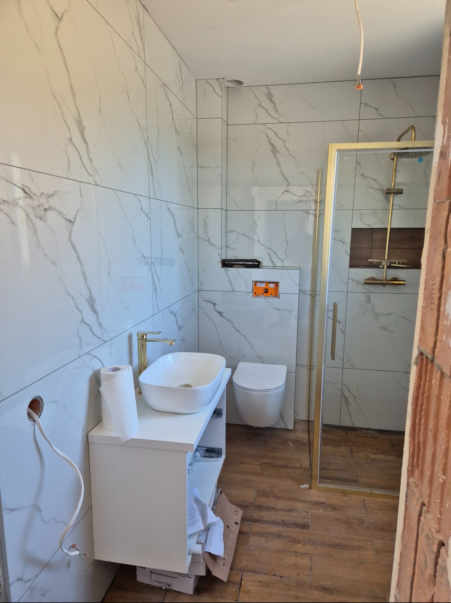 Biała łazienka z marmurowymi płytkami, złotą armaturą i kabiną prysznicową. Widoczne niedokończone prace: wystające kable, brakujące elementy wykończenia.